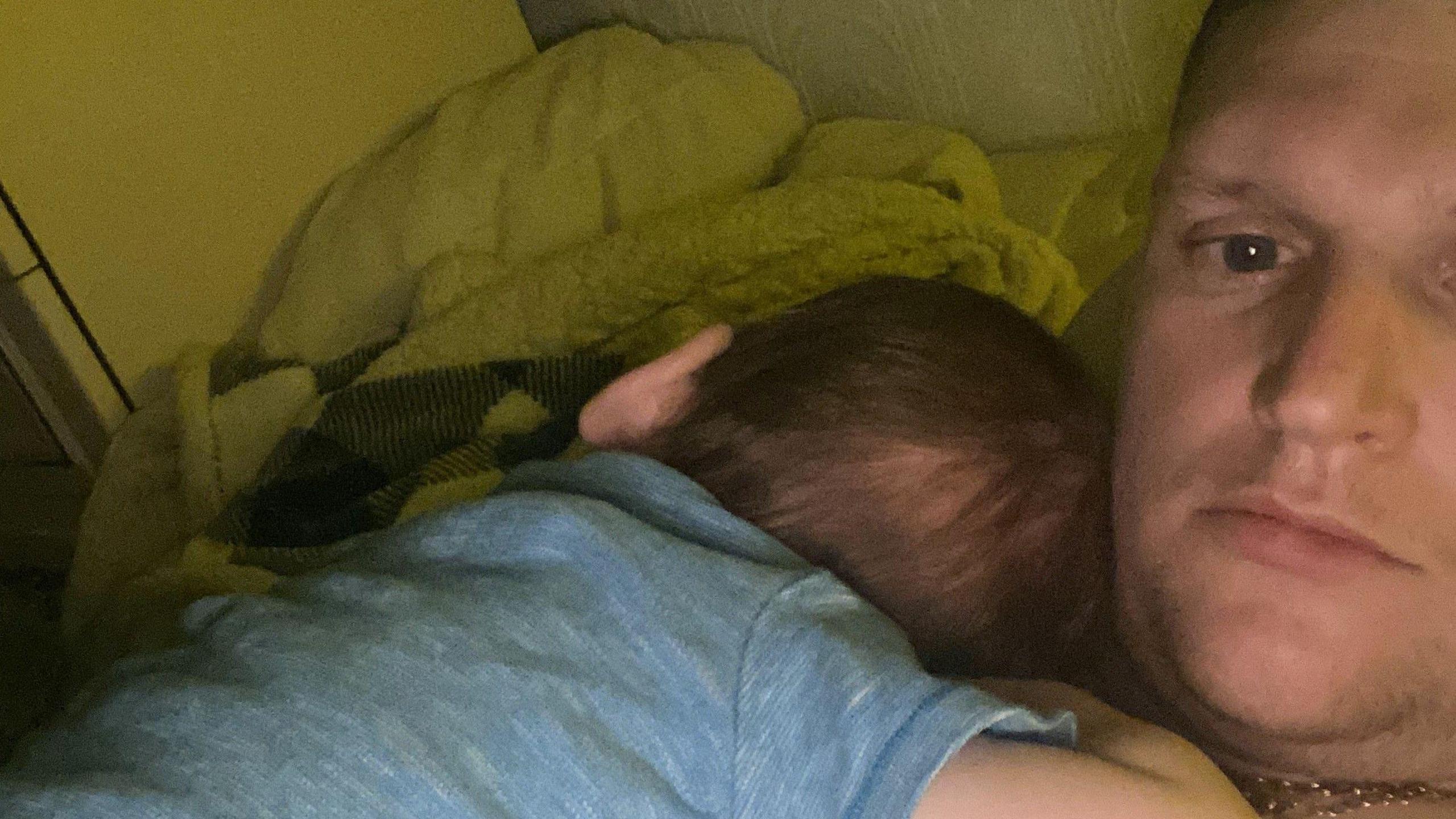 Charlie in a blue shirt is lying on Christopher Stockton's chest in a selfie photo. The child who has dark hair appears to be asleep.