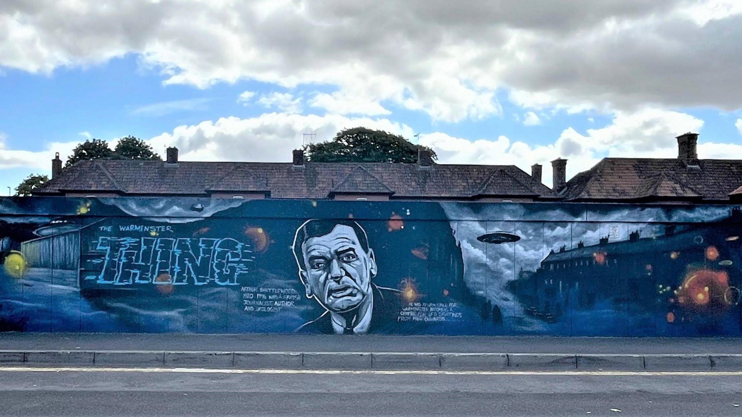 A wall painted with a mostly dark blue mural, with roofs of houses behind the wall. The mural reads 'The Warminster Thing' and includes a flying saucer near a row of terraced houses. A portrait of journalist and ufologist Arthur Shuttlewood is included prominently.