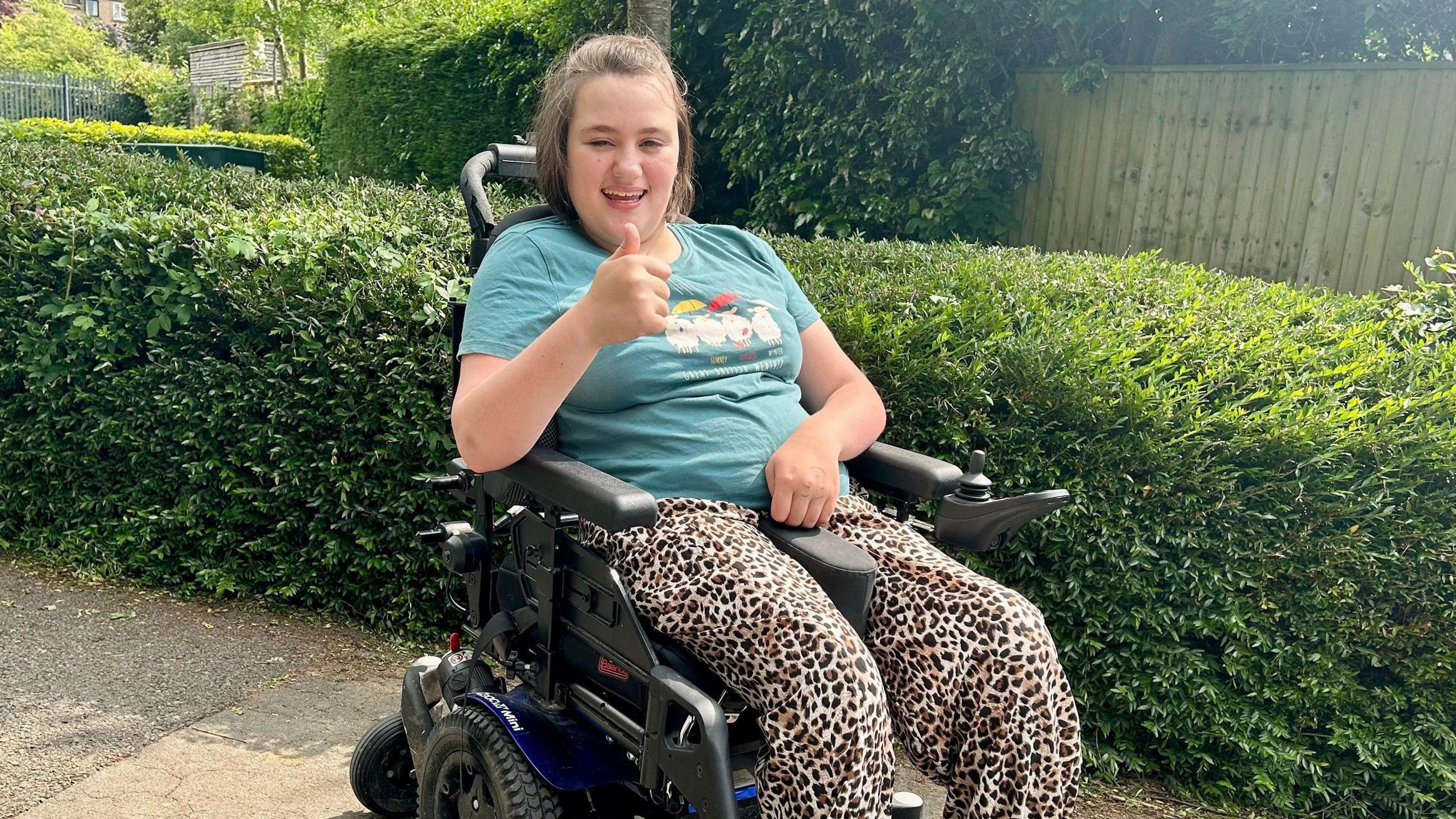 Charlotte pictured in a motorised wheelchair in front of a long green hedge in a garden, She is smiling and giving a thumbs-up, and wears a teal-coloured t-shire and loose leopard-print trousers. 