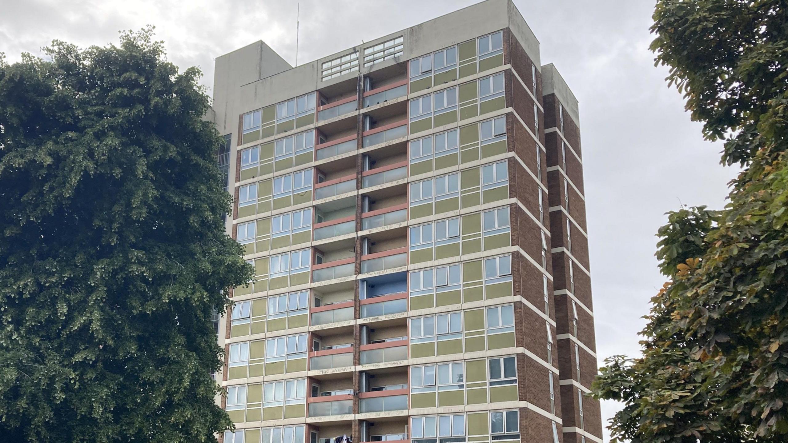 A photo of an 11-story tower block. There are many windows which are surrounded by green cladding. There are also many balconies with red cladding around. There are green trees either side of the building