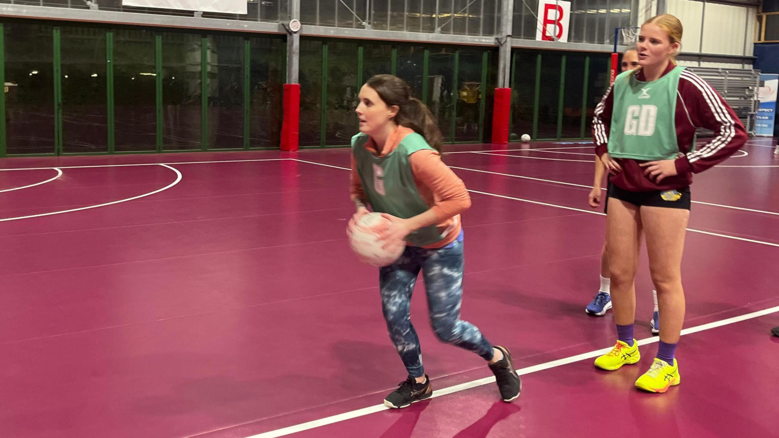 Sammy is holding a netball in both hands and is getting ready to pass it. Players are lining up behind her and she's looking at where she's going to pass the ball. She's wearing dark blue camo leggings with black trainers. 