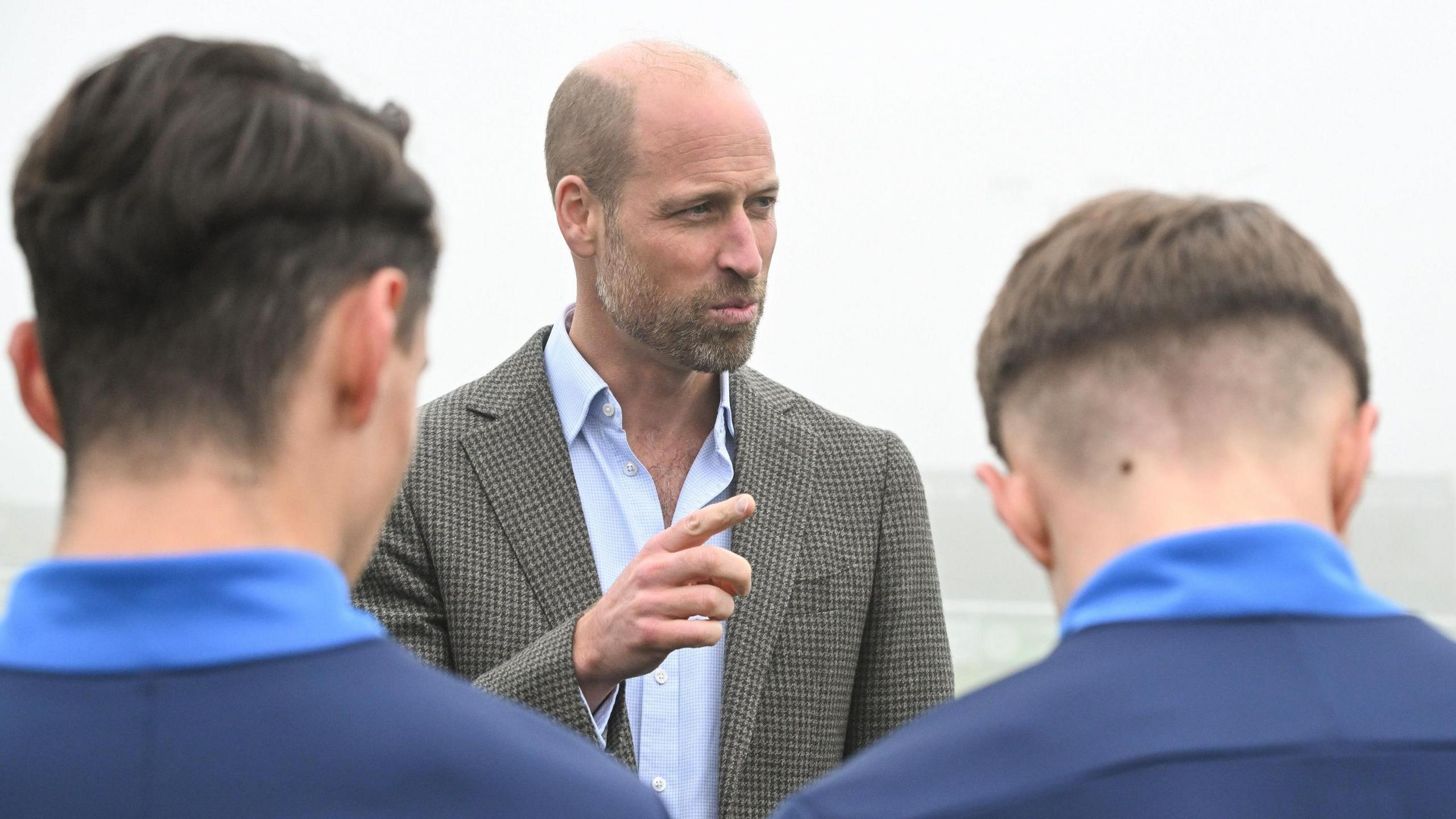 Price William is pointing a finger and two young men are looking at him with their backs to the camera. Prince William is wearing a pale blue shirt and a checked jacket. He has pale grey hair and a beard. It is a foggy day.