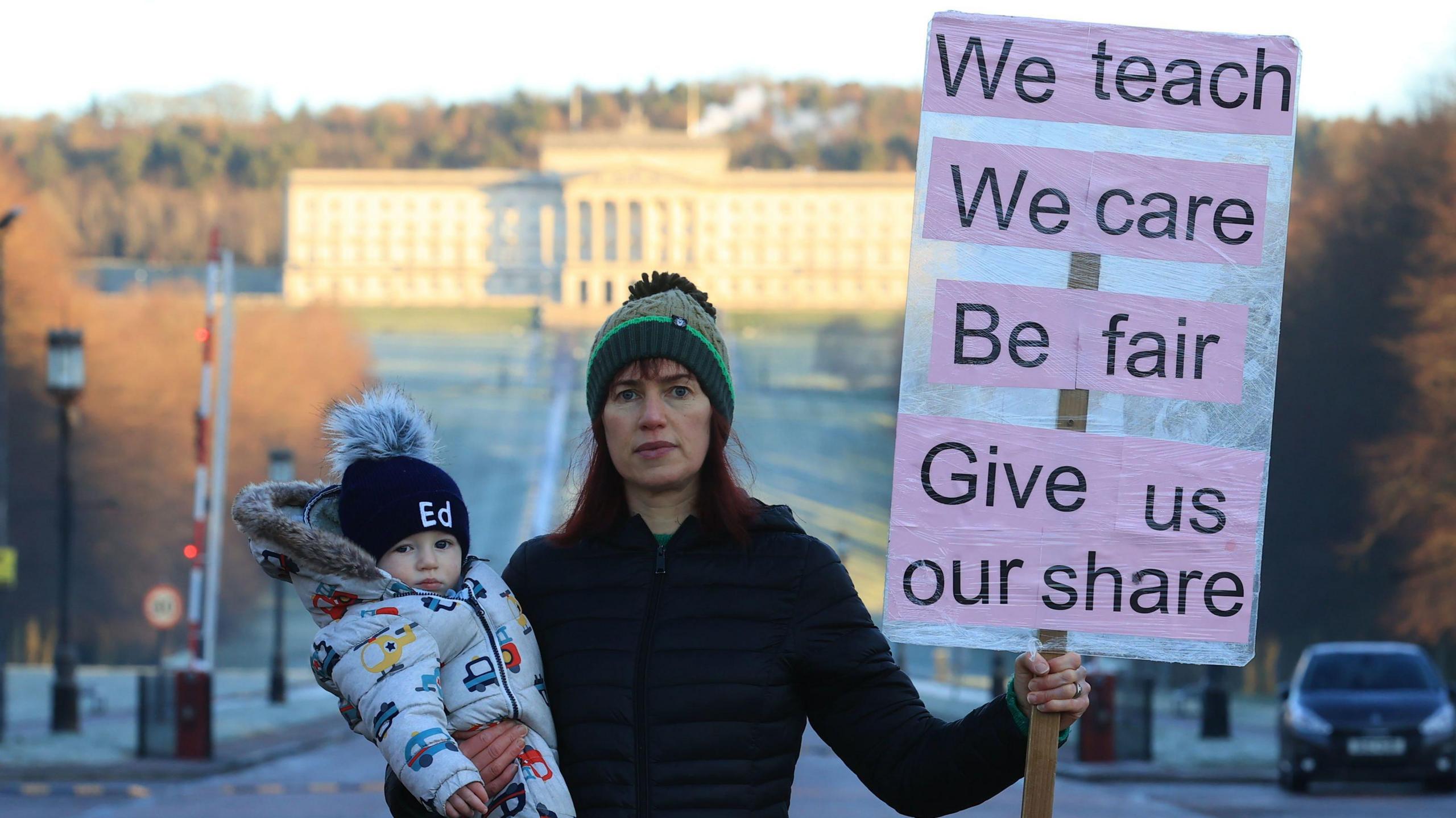 A teacher dressed in a bobble hat and a dark coat, stands holding her baby in one hand, and a placard reading "We teach. We Care. Be Fair. Give us our share". It is a cold frosty morning and parliament buildings at Stormont are in the background.