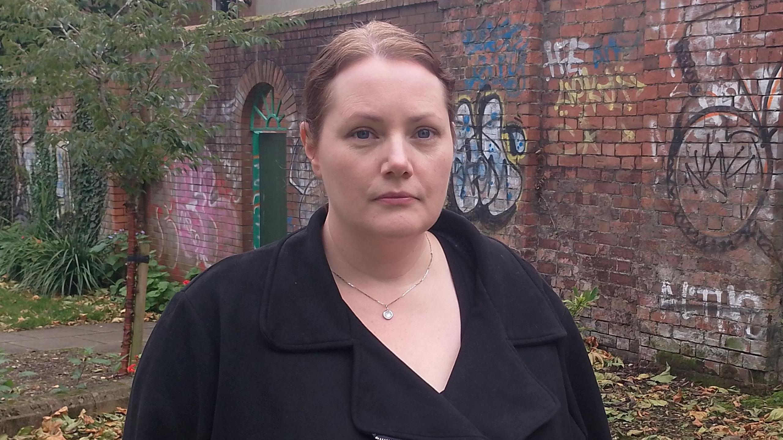 Elaine Crory from the Women's Resource and Development Agency. She is standing in front of a red brick wall which is covered in graffiti. She had light brown hair, which is tied up. She is looking at the camera with a neutral expression. She is wearing a silver necklace, and a black coat.