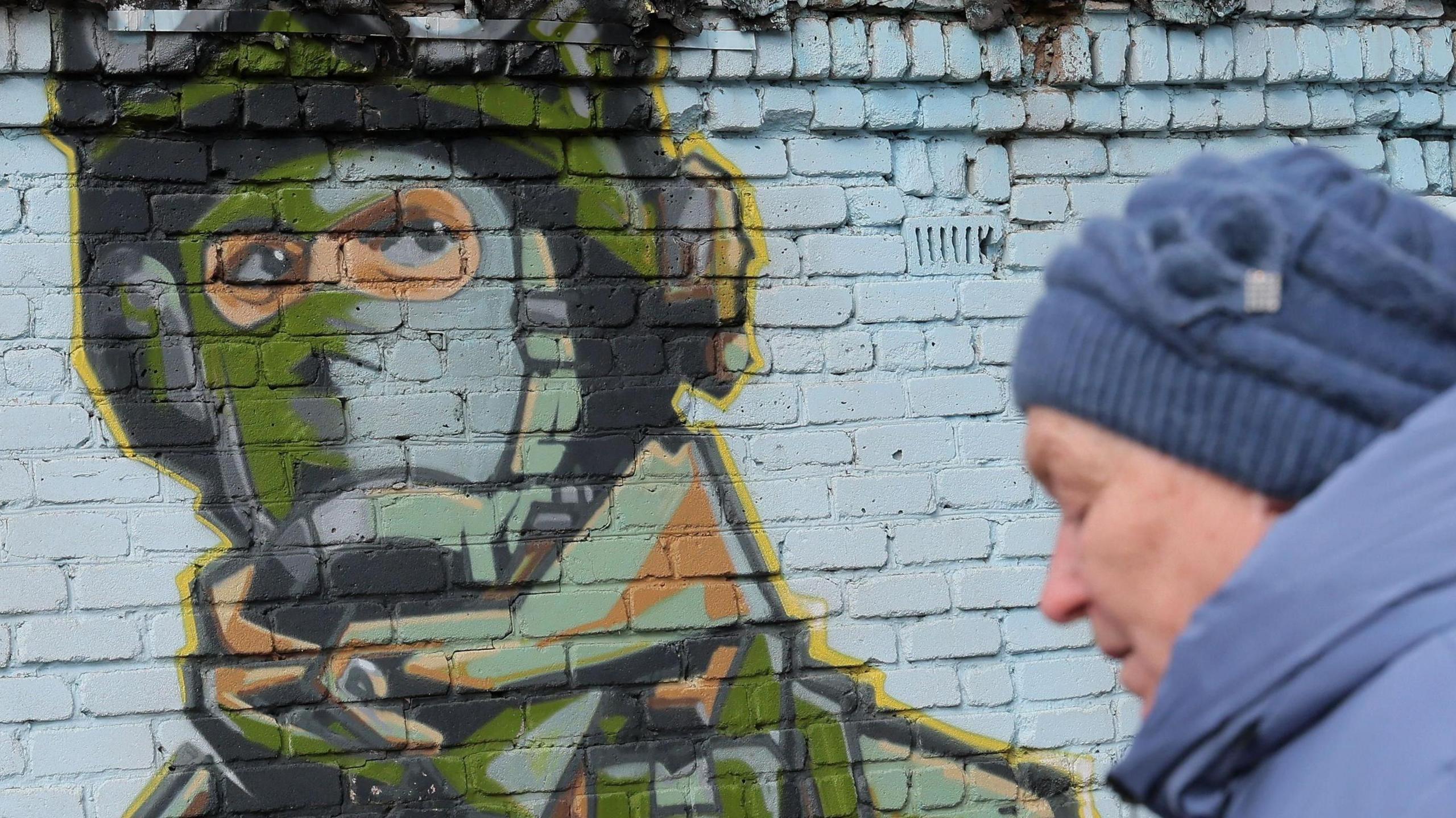 A woman walks in front of graffiti on a wall showing a Russian soldier 