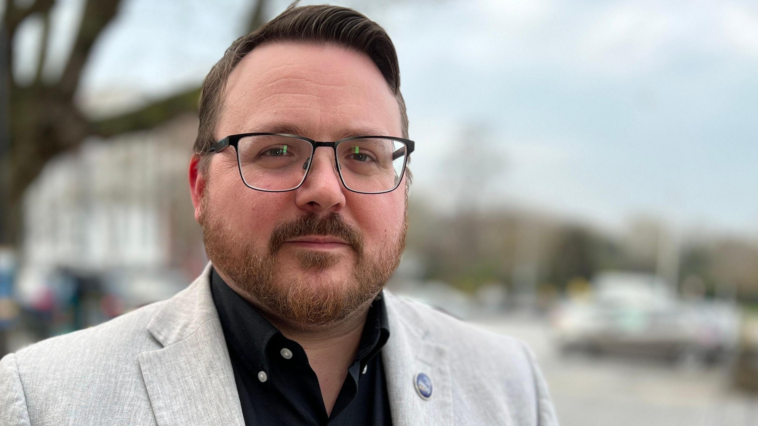 Daniel Cowan is seen on the left hand side of the screen. He has brown hair, a brown and ginger beard, and it wearing glasses, looking towards the camera. 