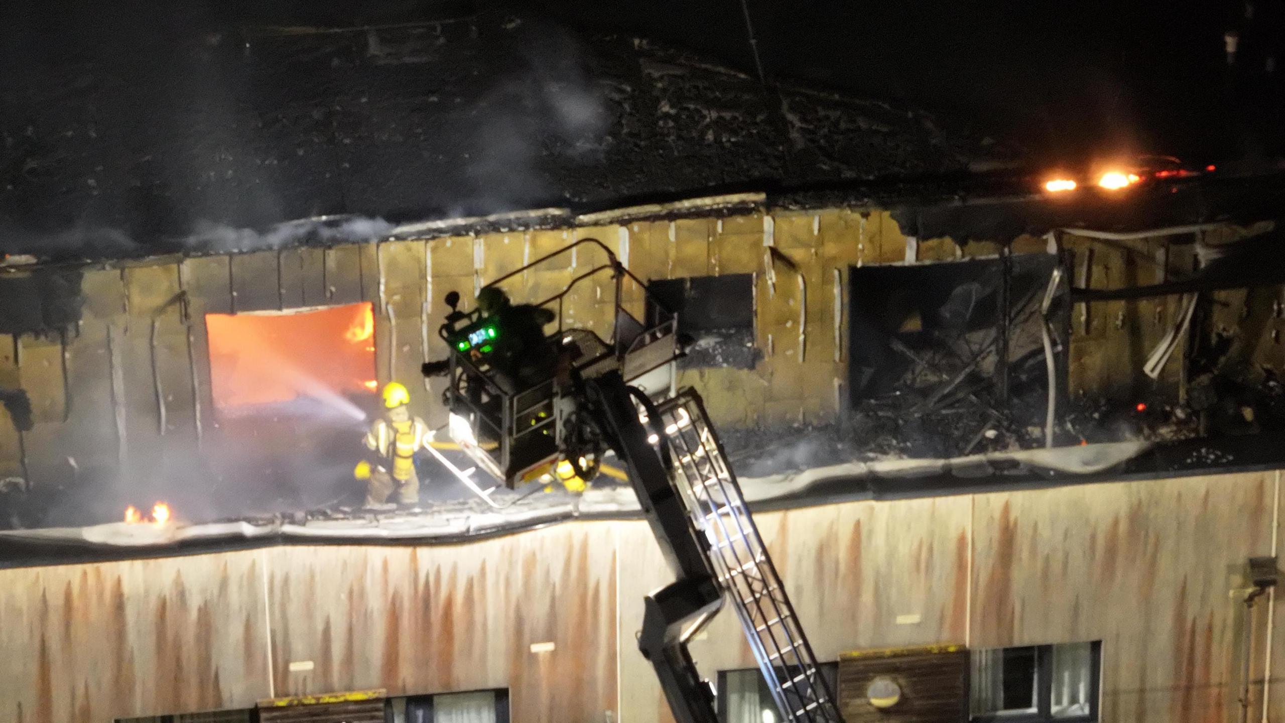 An aerial view of a firefighter on a balcony spraying water through a window on to the fire