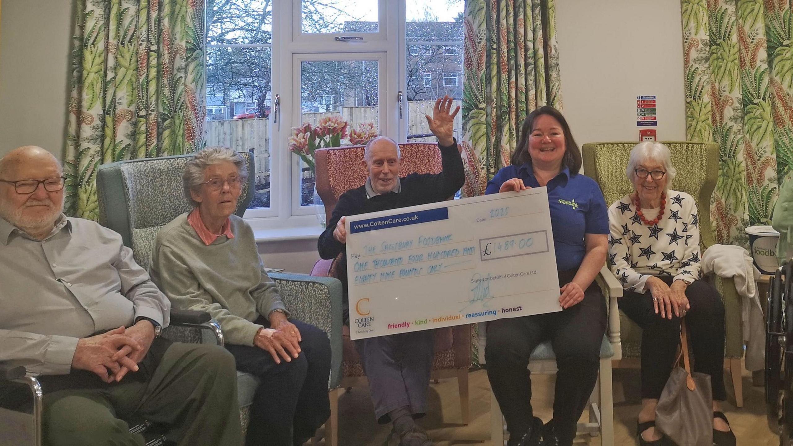 A group of elderly care home residents sitting inside - one sits next to a staff member as they hold a large charity cheque.