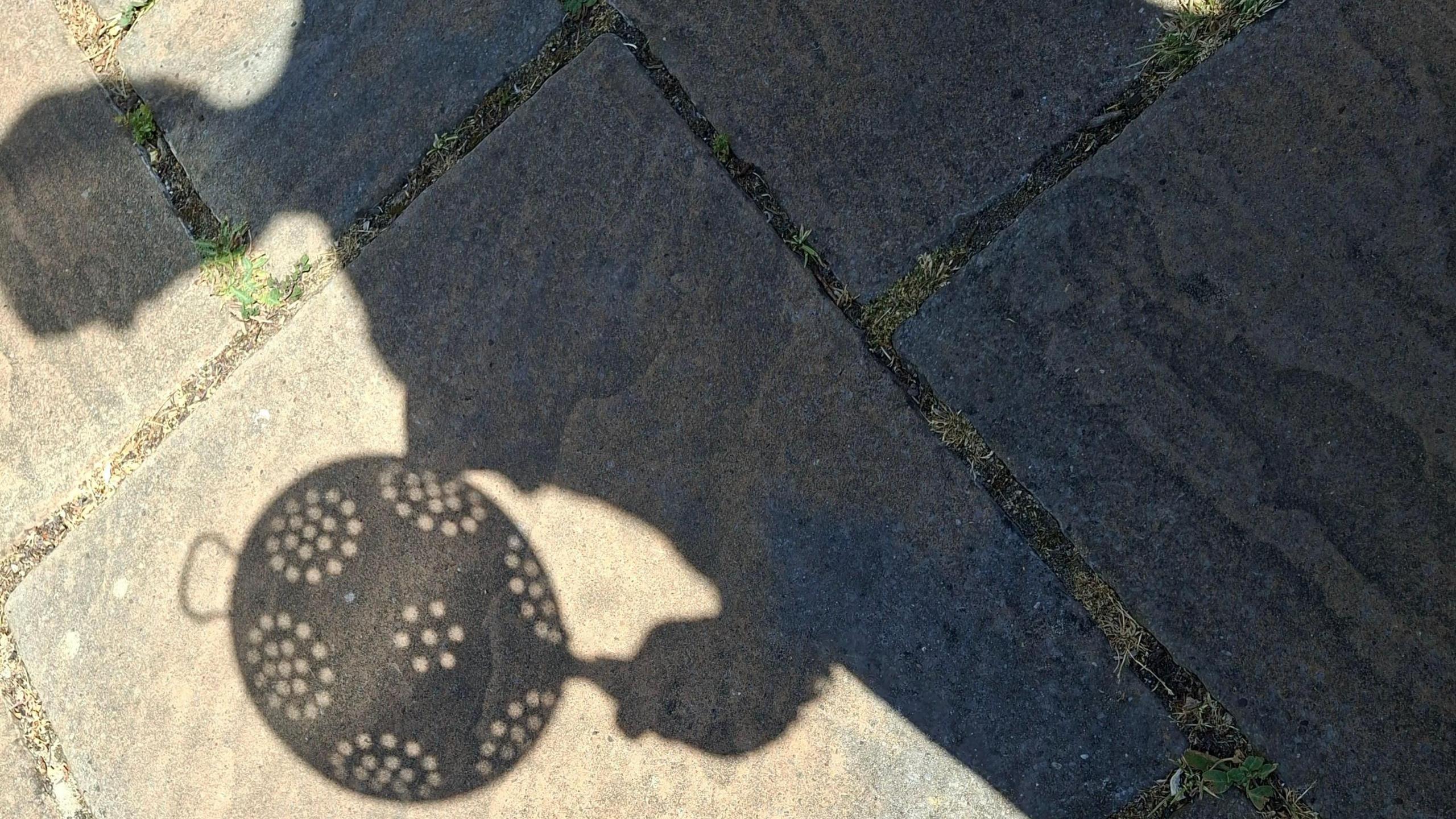 The shadow of a person holding a colander falls across paving stones in bright sunshine in Great Bookham, Surrey. The outline of the colander and its holes can be seen clearly on the ground.