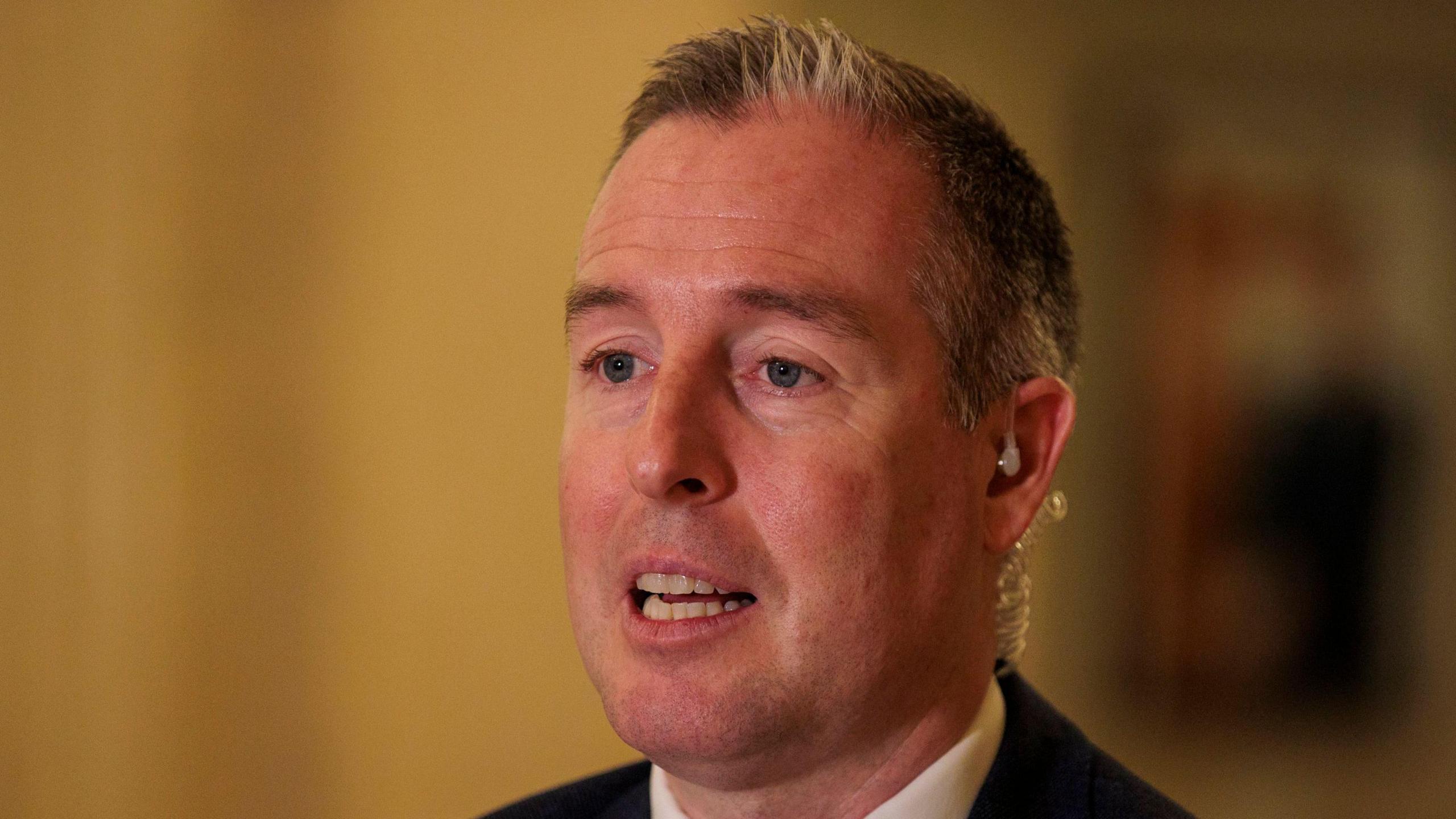 Paul Givan at Stormont Hall, he has short white hair and is wearing a blue suit with a red tie and white shirt. He is speaking and has an earpiece in. 
