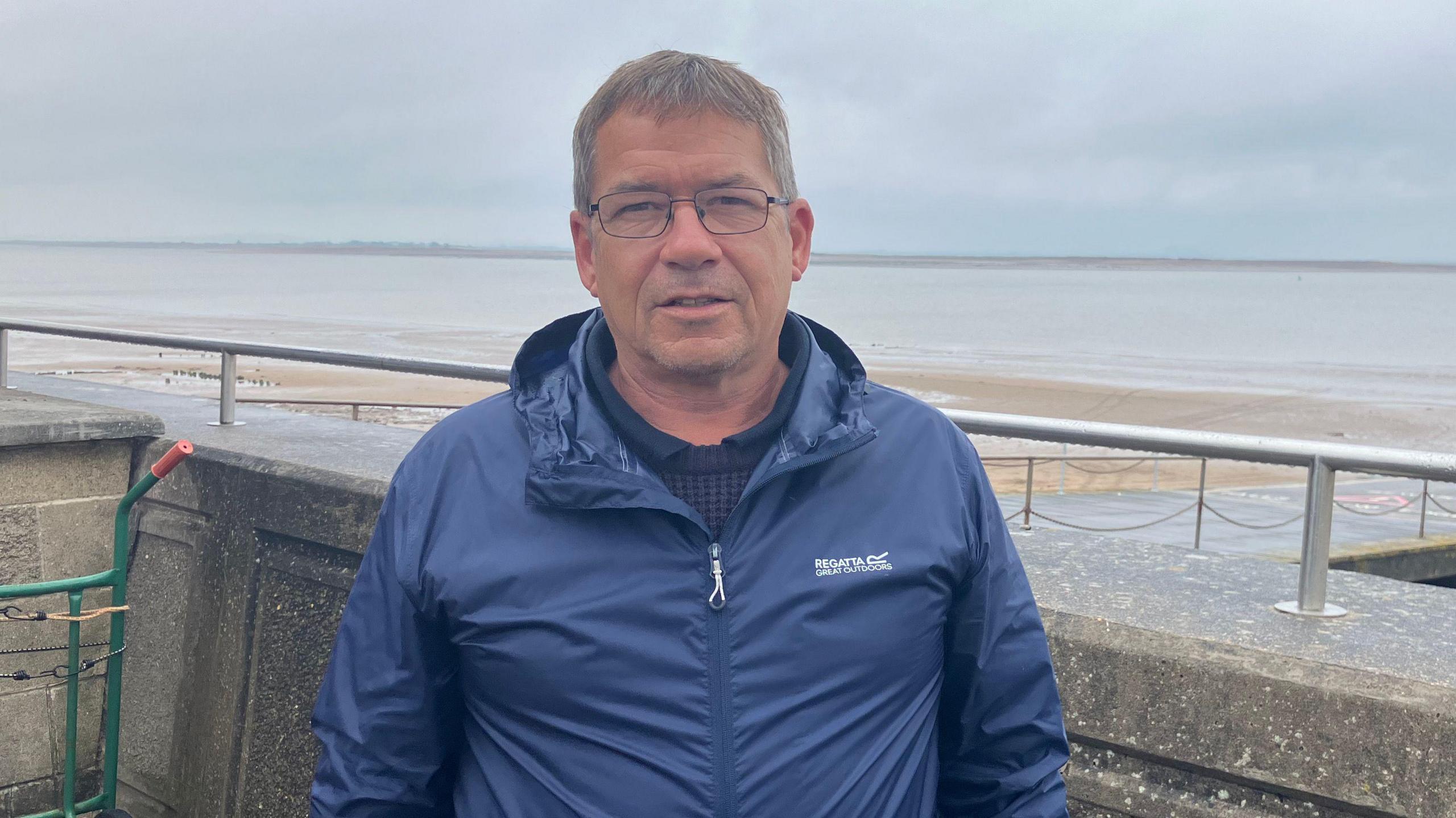 Ian Jeffries standing on the coast looking at the camera. He is wearing a blue jacket and the skies overhead are cloudy