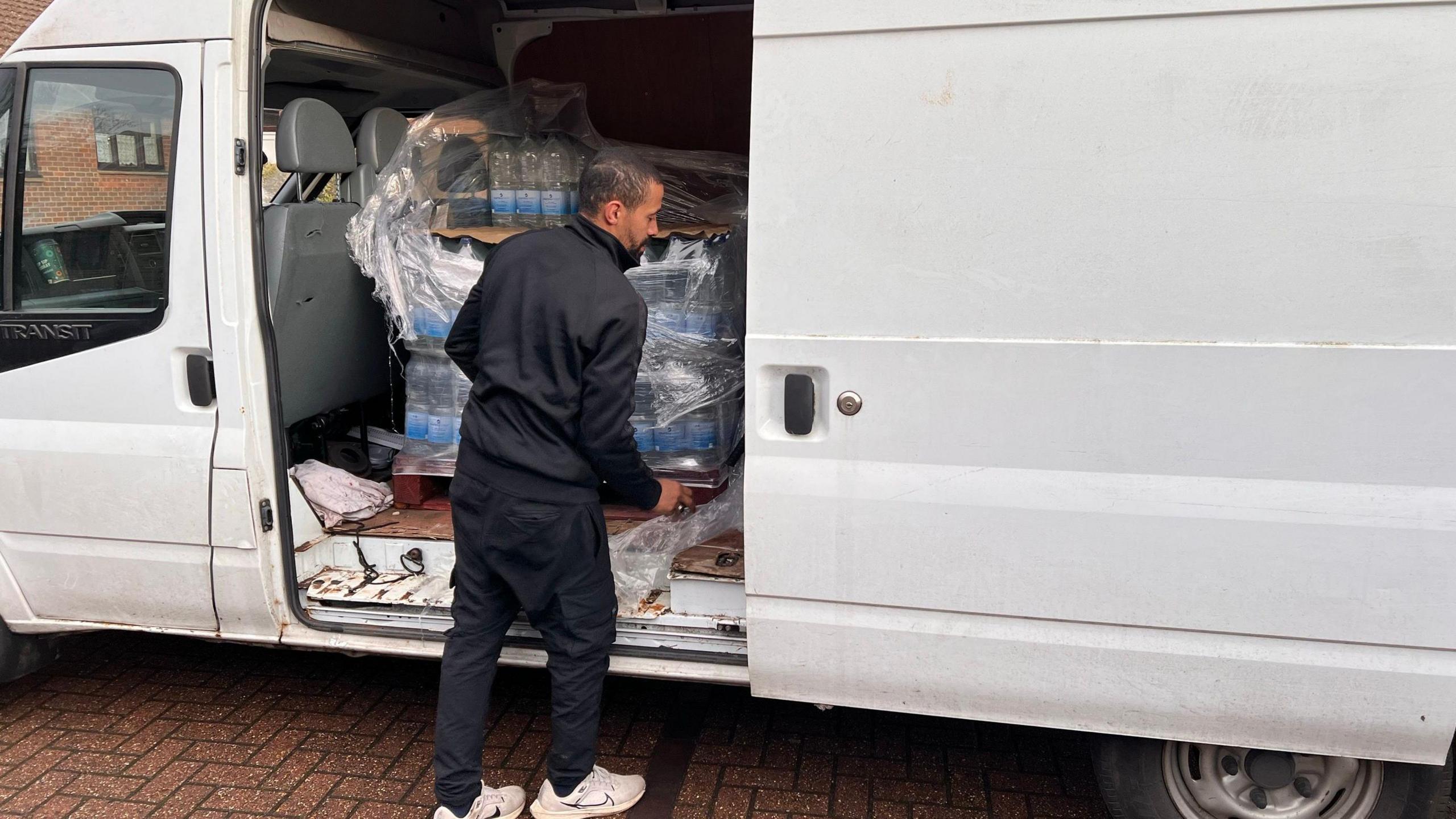 A man unloads pallets of water from a white van. The man is wearing a black top and black trousers. 