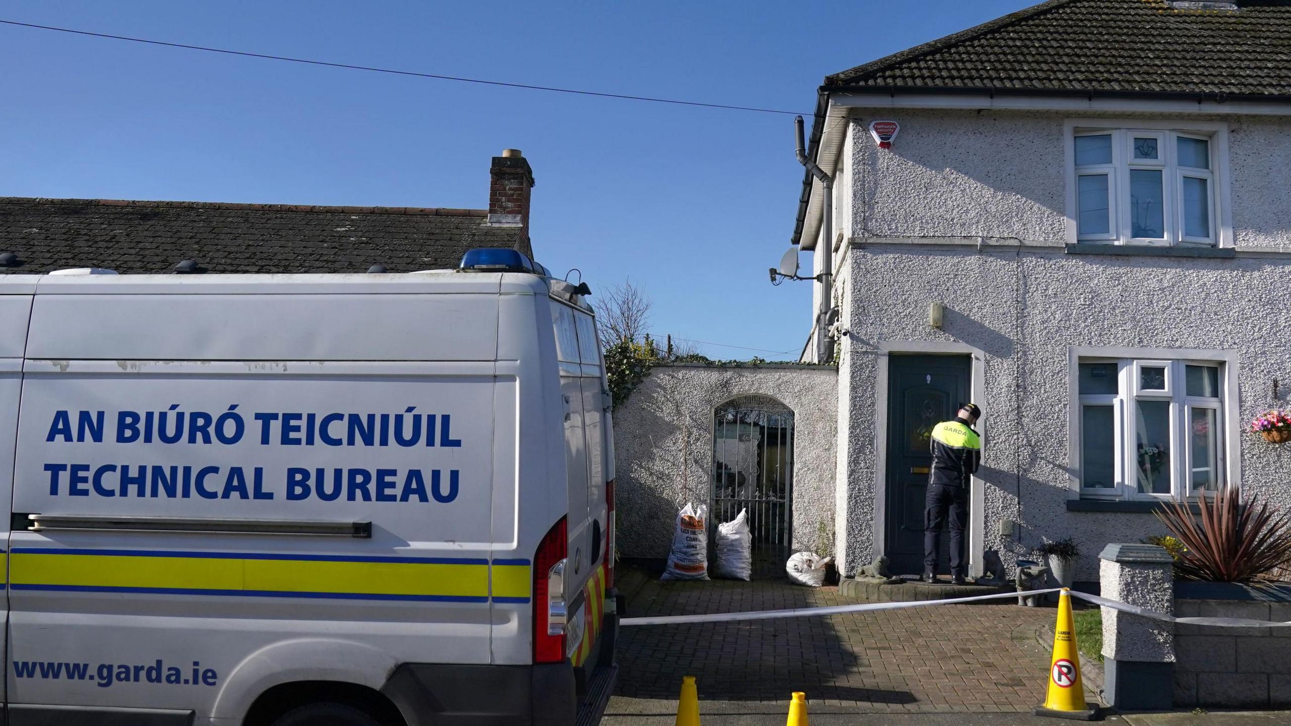 A white terrace house with a brown roof and dark coloured door. A gardaí officer is standing at the door of the house wearing police uniform which is black trousers with a black and yellow hi-vis top. Outside the house is yellow cones and tape to create a cordon. A white 'technical bureau' van with blue and yellow detailing is parked outside.