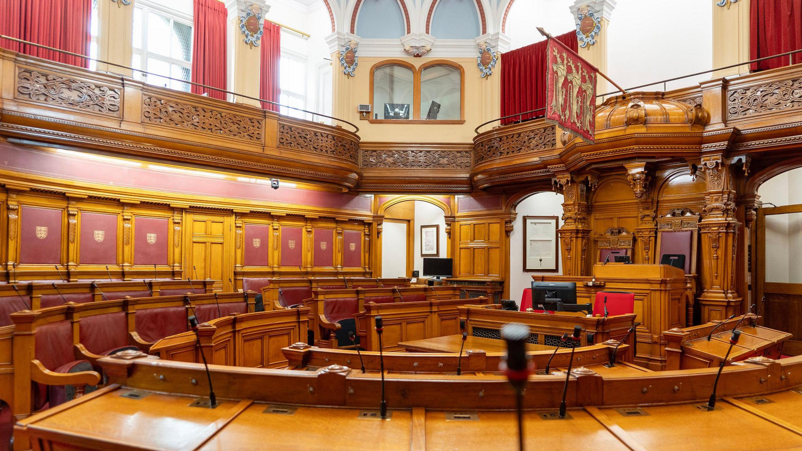 The interior of the States Assembly which is a round room lined with wooden benches with red leather backs that have desks in front of them. The second storey has windows with floor-length red curtains and the seat the Presiding Officer sits in features wooden carvings and has the Jersey flag hanging above it. 