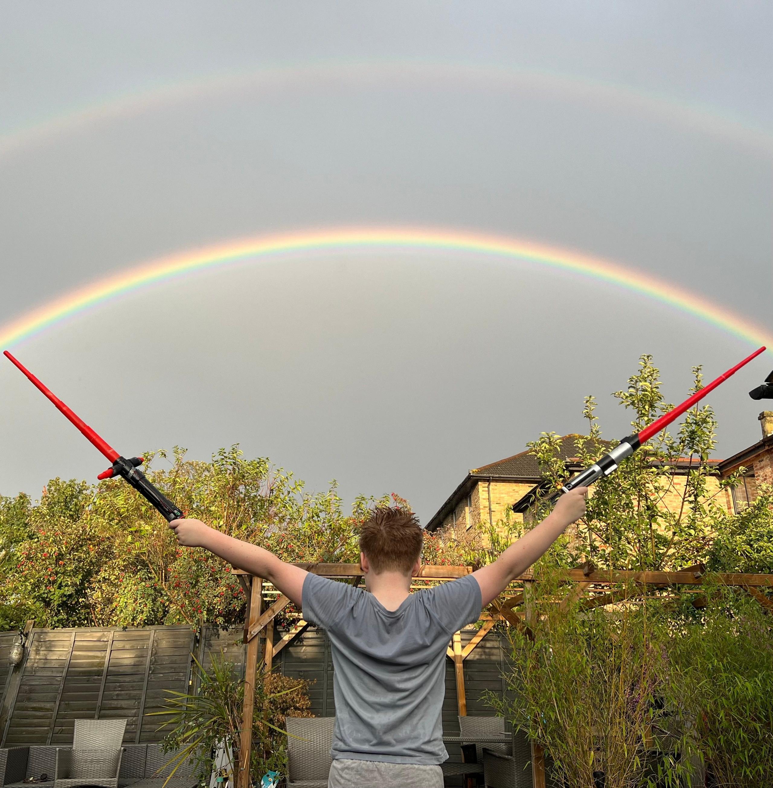 A boy holds two lightsabres at either end of a rainbow