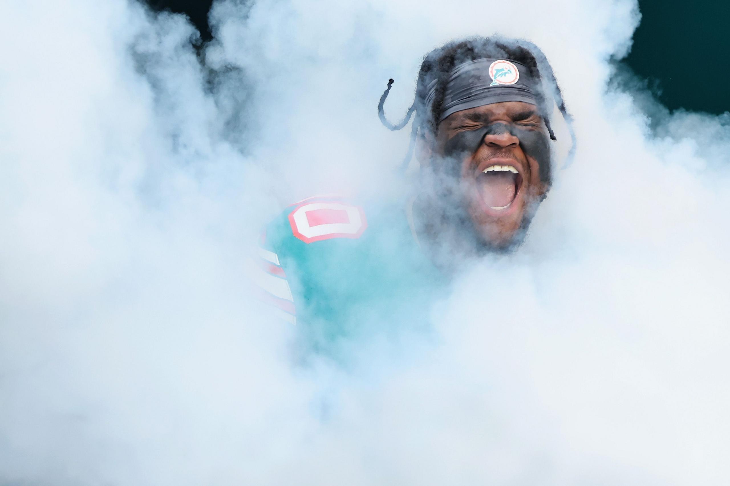 An American football player wearing a teal jersey with the number 10 and a helmet featuring a dolphin logo emerges through thick white smoke, creating a dramatic entrance scene. The smoke surrounds the player almost completely, with only parts of the jersey and helmet visible.