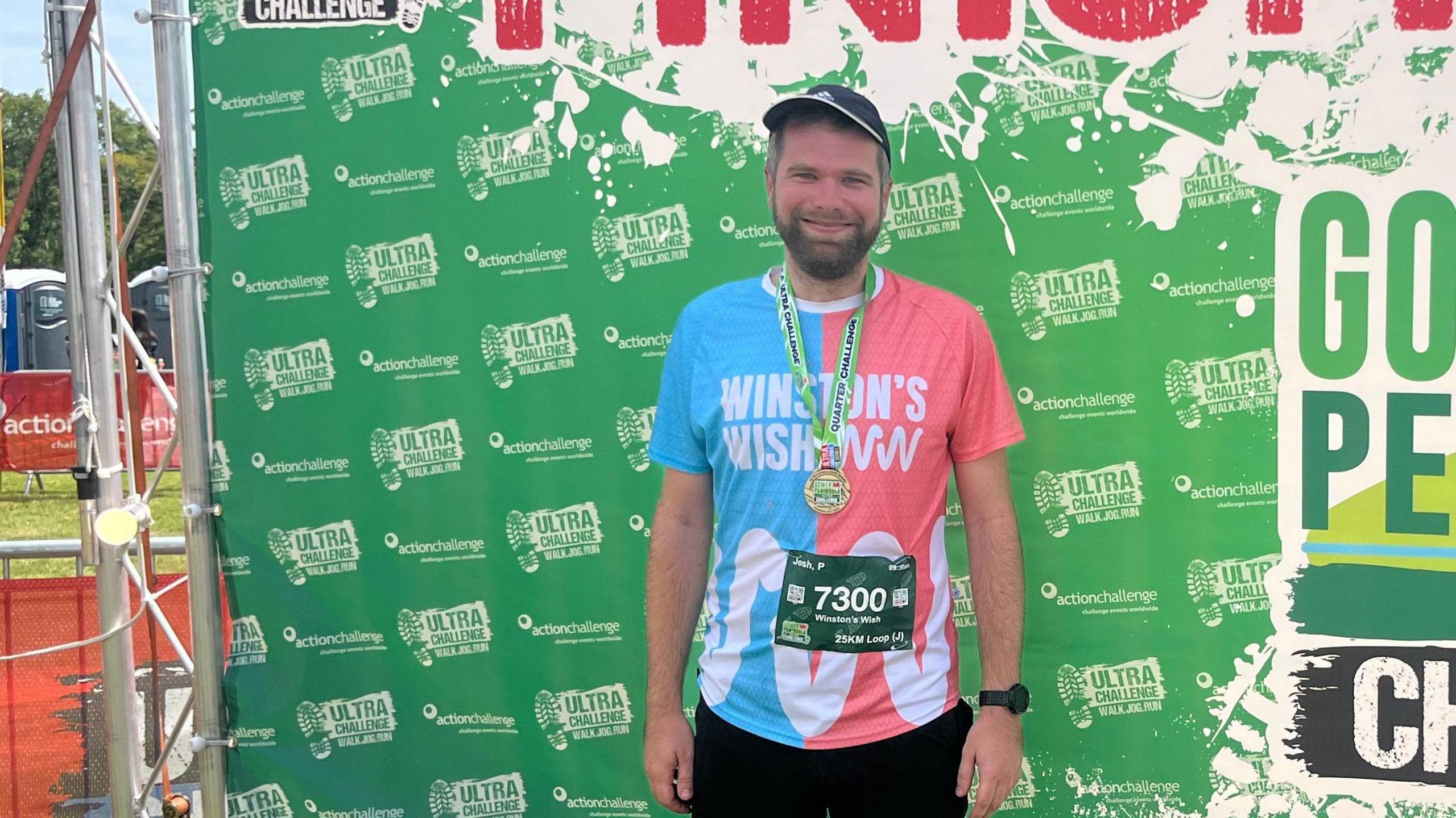 Josh Preece wears a pink and blue Winston's Wish sports t-shirt and a green and gold medal. He is smiling in front of a green sign at the end of a race. 