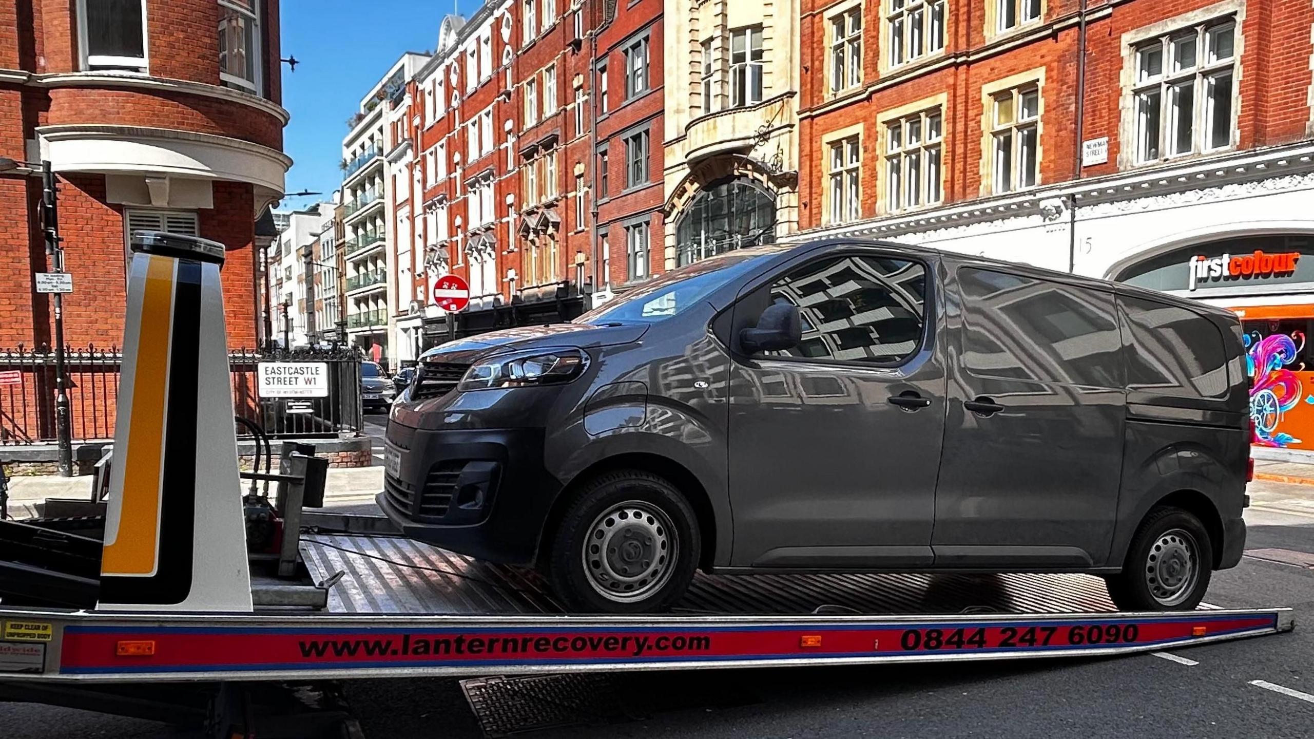 A grey van is loaded onto a recovery truck on a street in London, with red brick buildings and a print shop called "first colour" visible in the background.
