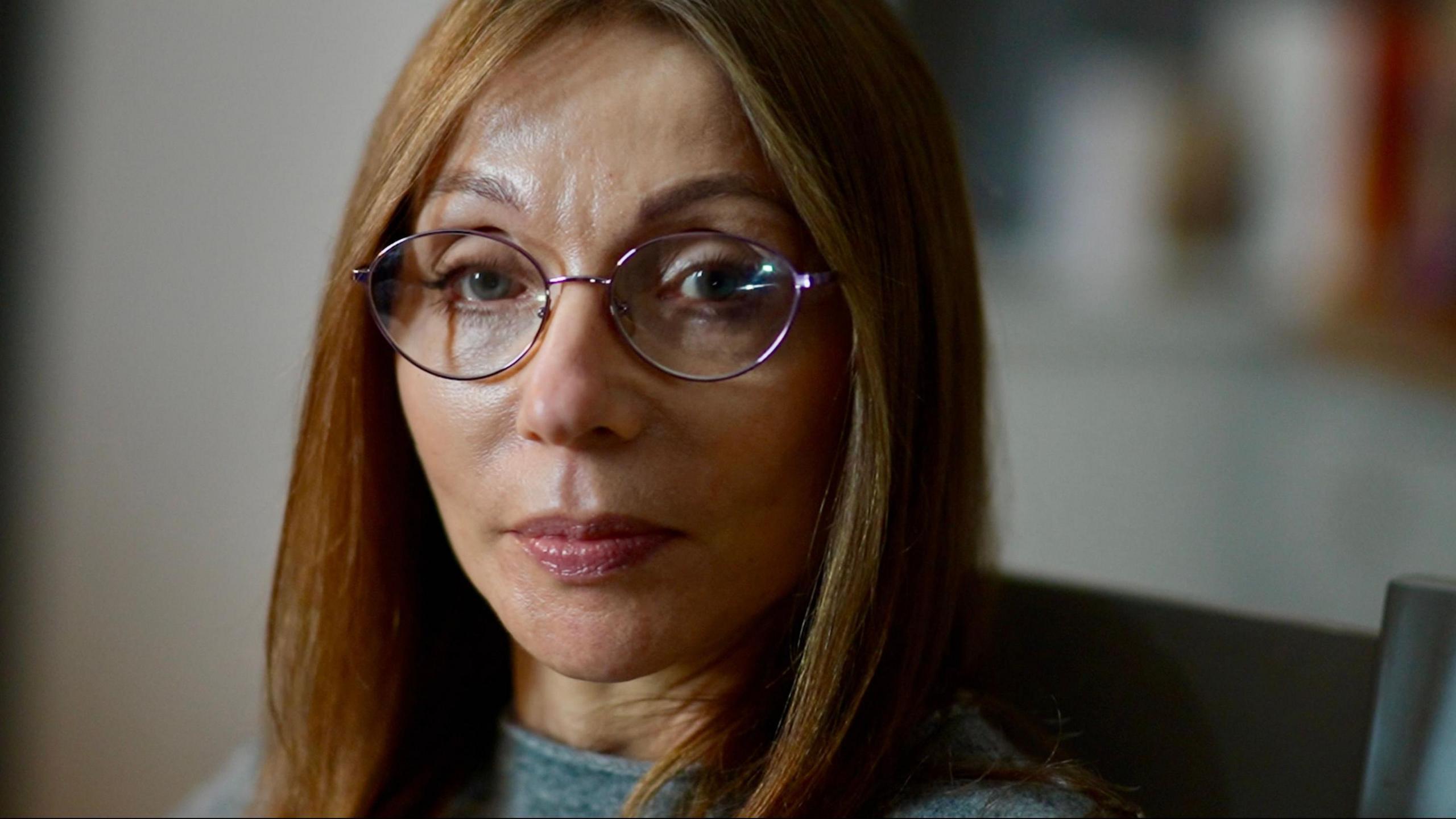Sharon, Amy's mother looks at the camera lens. She is framed slightly to the left of the picture and has light brown to dark blonde, long, straight hair. She has chrome-framed rounded glasses on and is wearing light make-up. She has a light grey wool jumper on and the very top of her shoulders are in the frame.