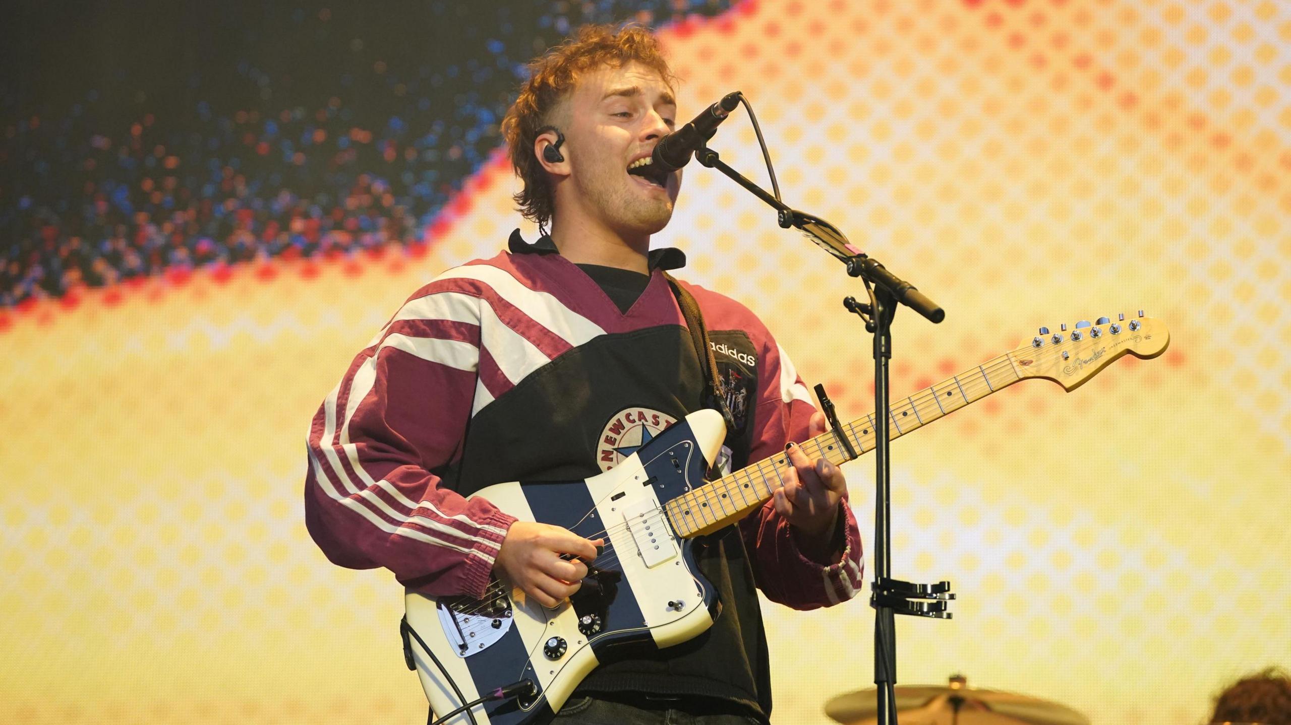 Sam Fender singing on stage. He his holding a black and white guitar and wearing a Newcastle United top.