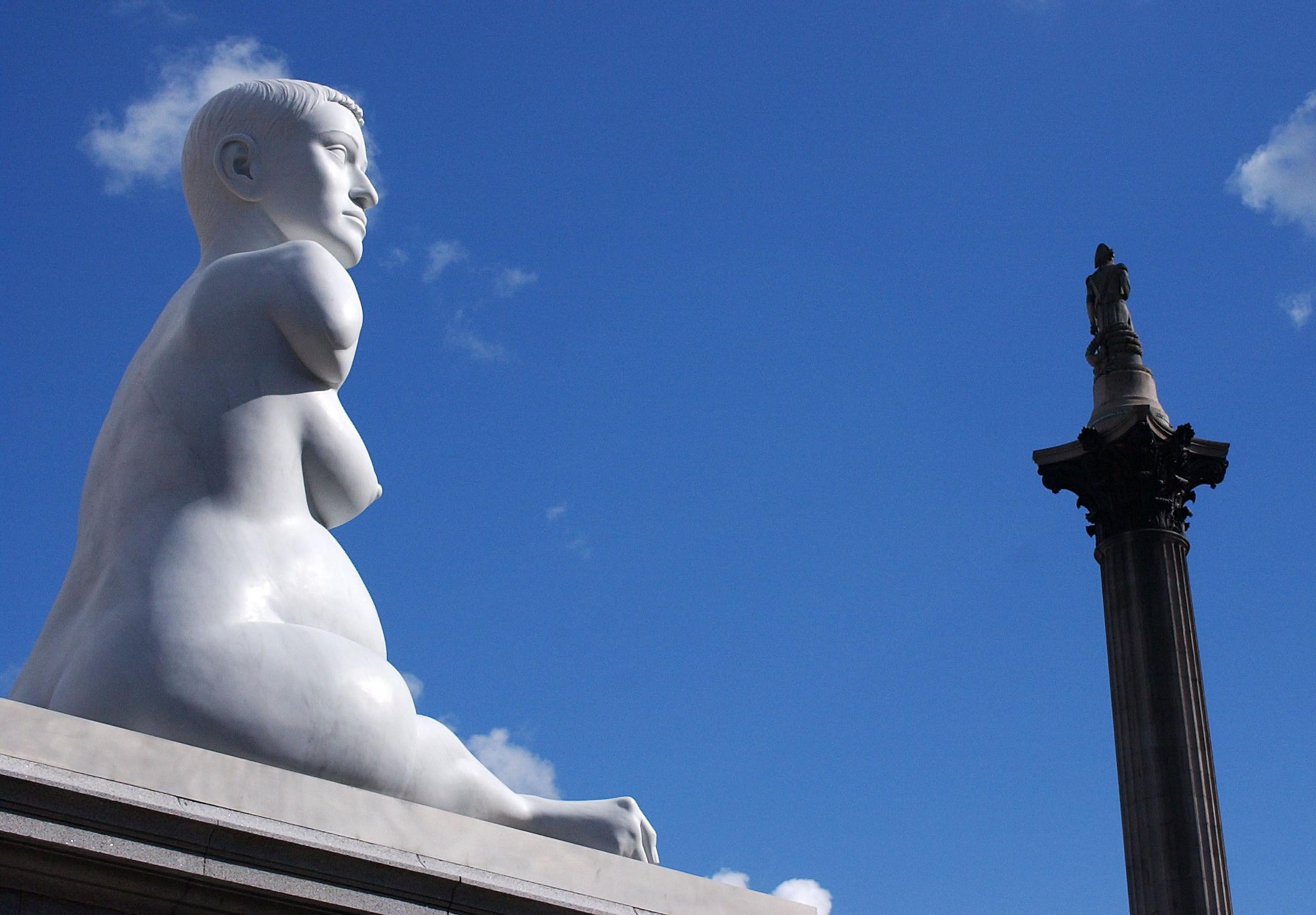 The sculpture, Alison Lapper Pregnant, in London's Trafalgar Square. The sculpture sits on a plinth. In the distance Nelson's column can be seen. At the top of the column is a statue of Lord Nelson. The sky is bright blue.