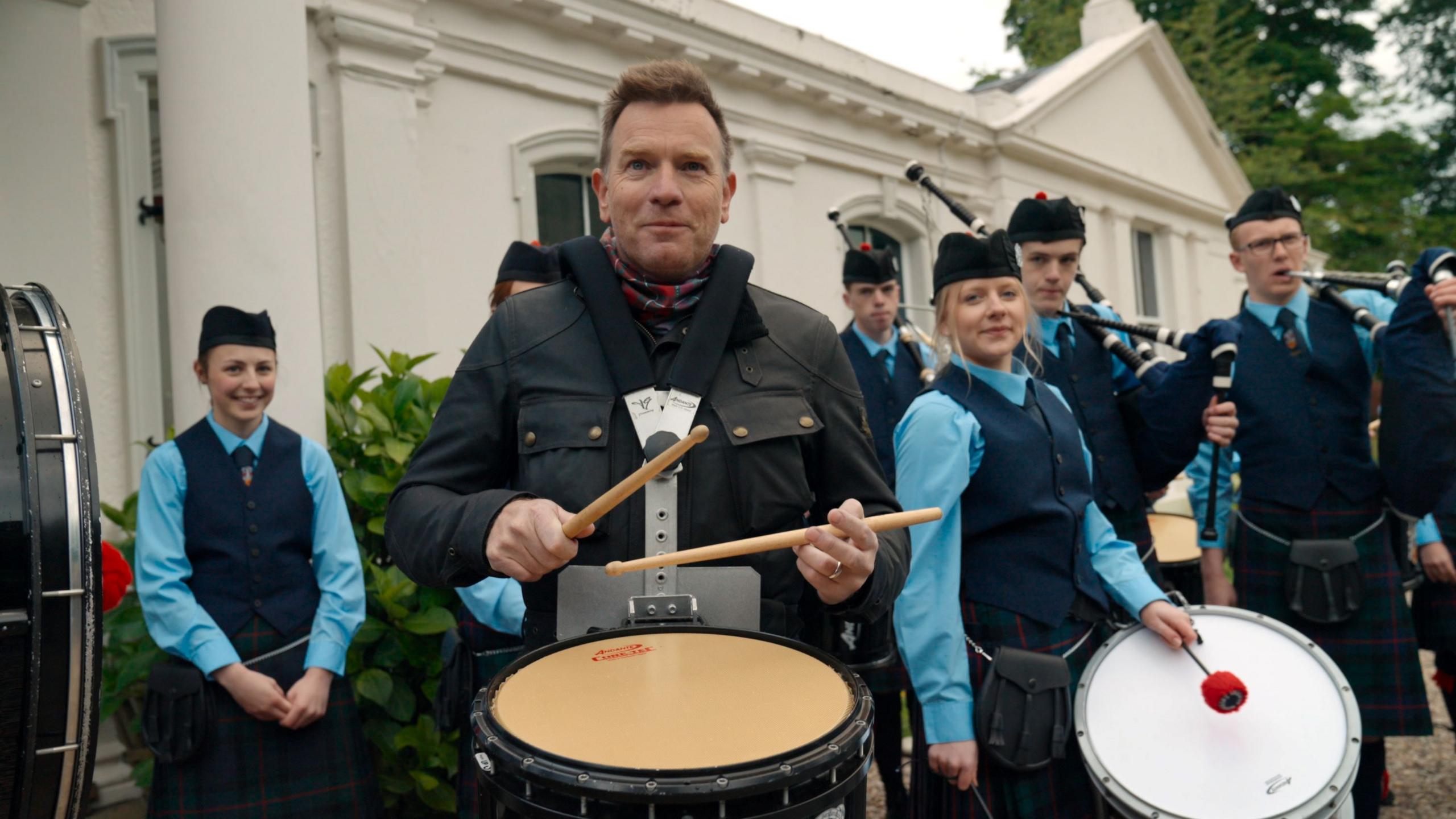Ewan McGregor, in a dark oilskin jacket, plays a side drum as members of Morrison's Academy Pipe Band stand around him. They are wearing school uniforms of navy tank tops and pale blue shirts.