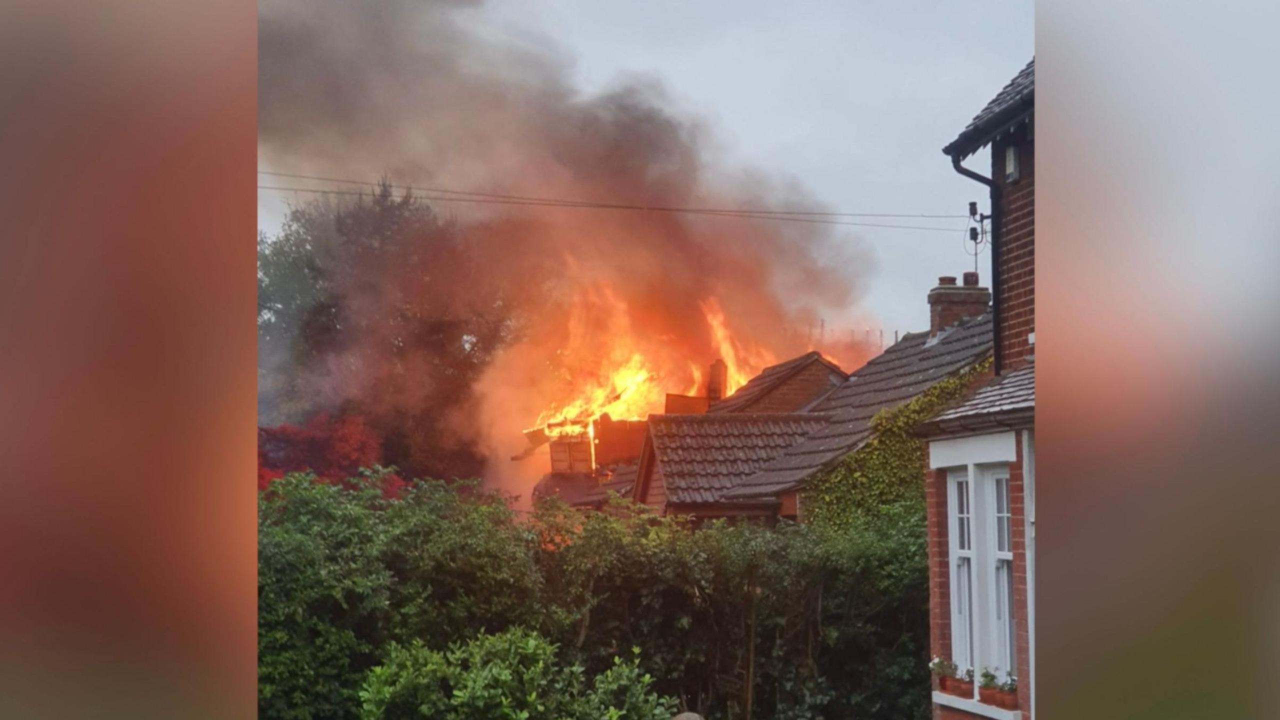 A photo shows a burning house with flames and smoke billowing from the top of it. 