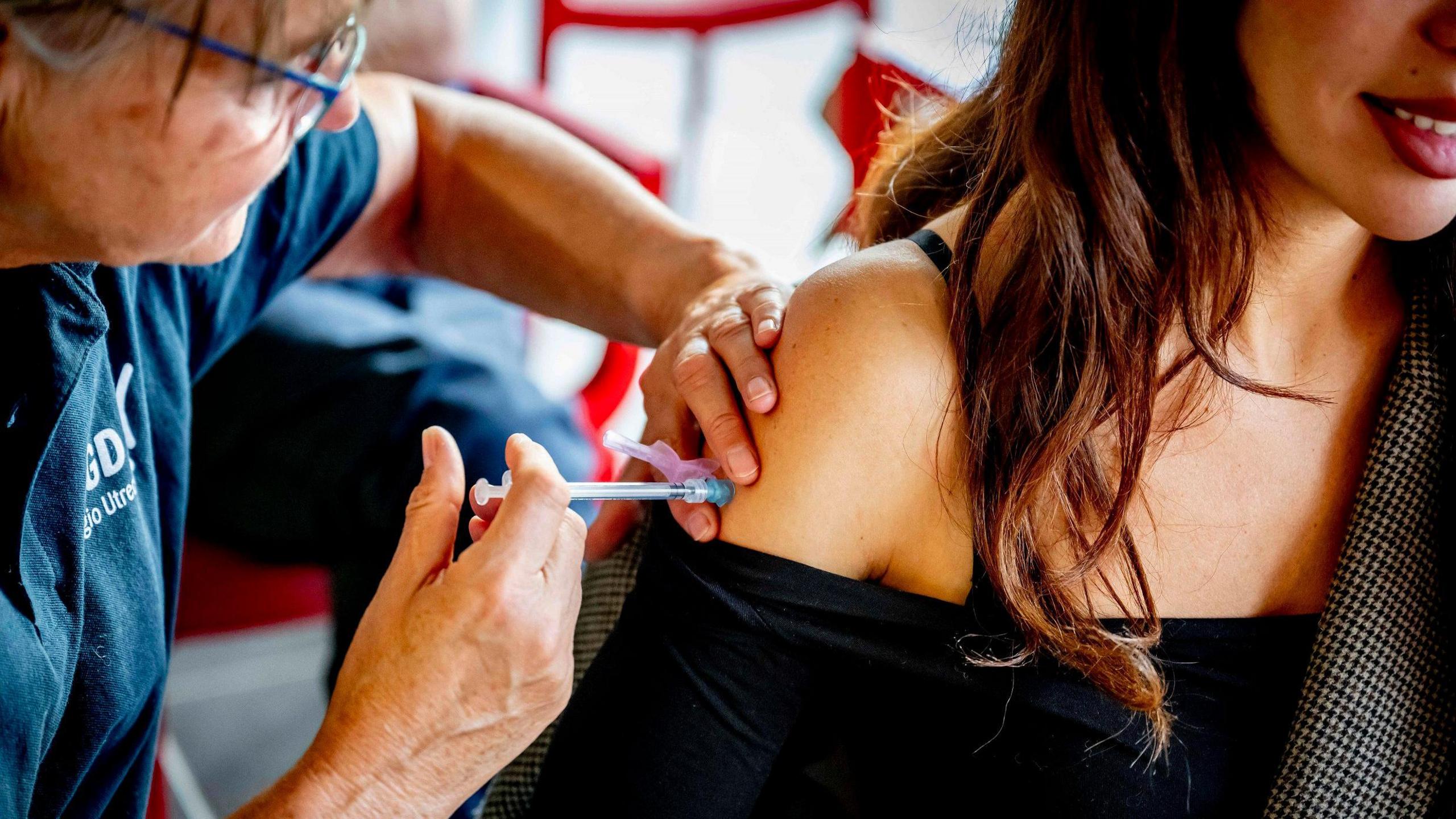 A woman is injecting a young woman. The nurse is wearing glasses and a T-shirt and has one hand holding the young woman's arm and the other is pressing the needle. The young woman has her shoulder bared for the injection and has long shiny brown hair and is smiling.