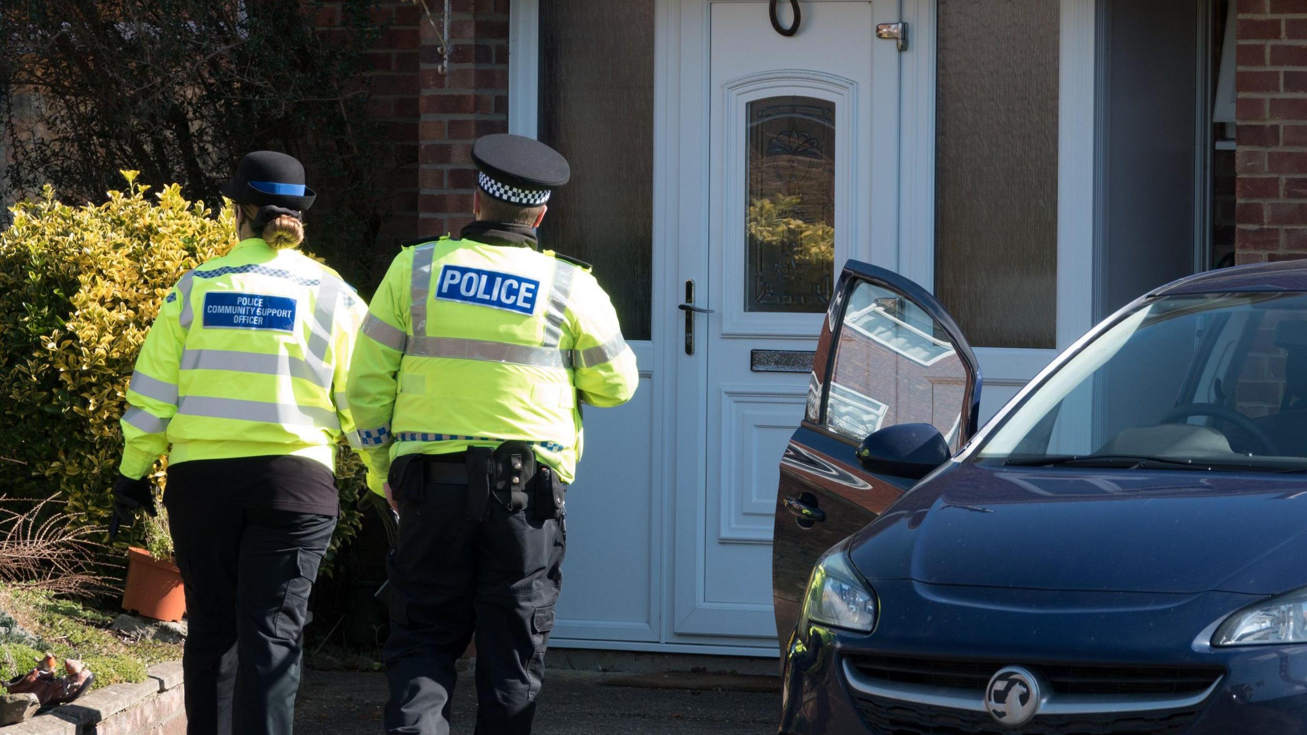 Two police officers, one male and one female, walk away from the camera towards the house of Sergei Skripal in Salisbury at the time of the Novichok poisonings. They are both wearing large yellow high-vis jackets. The house has a white front door and a dark blue Peugot car is parked outside