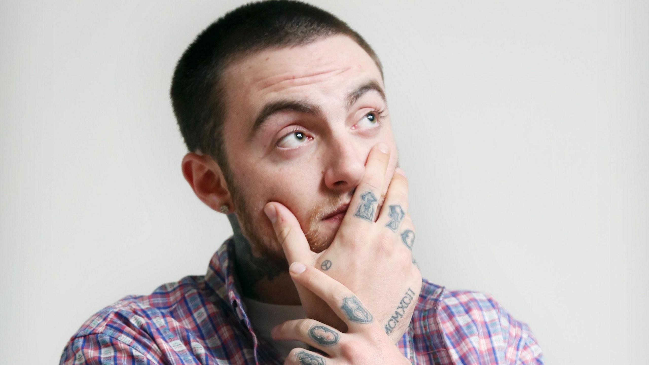 Mac Miller pictured with his tattooed hand covering his face and looking up away from the camera. He wears a blue, white and pink plaid shirt over a white T-shirt and is photographed against a bare white wall. 