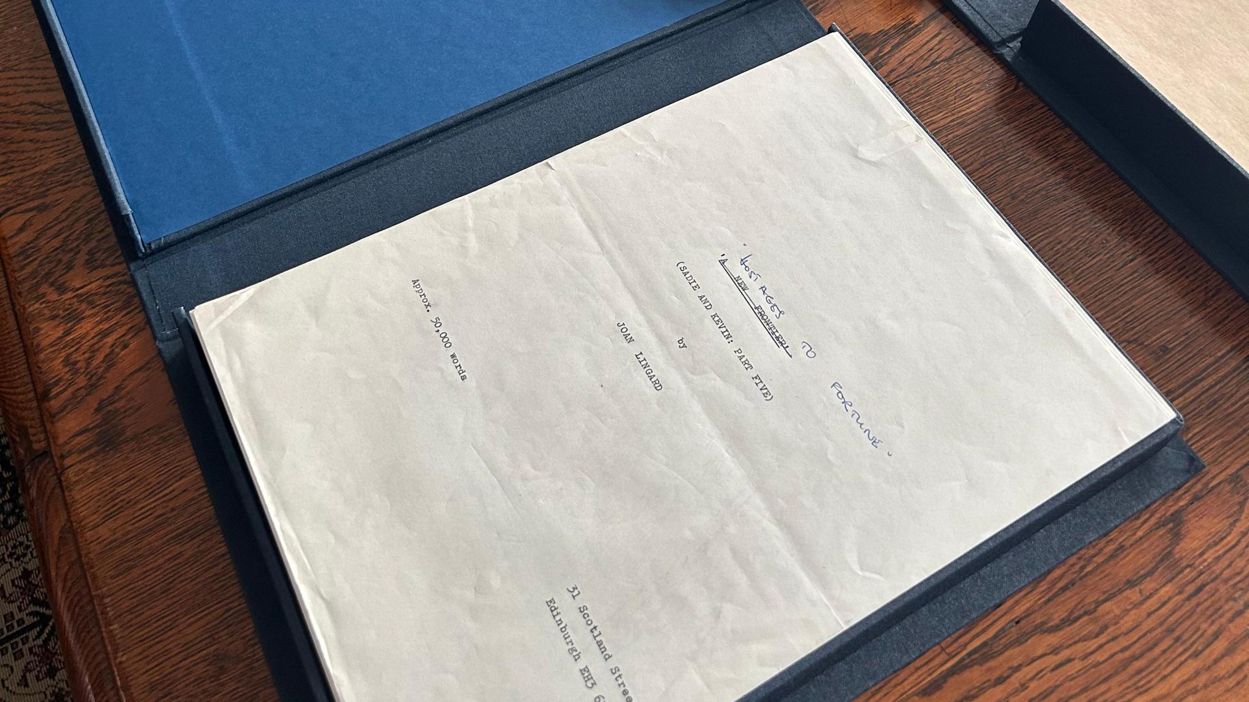 A navy folder is sitting open on a wooden desk. Inside it is a worn paper document with some typed words saying (SADIE AND KEVIN PART FIVE) BY JOAN LINGARD. The typed title has been scored out with a pen and the words "Hostages to Fortune" handwritten beside it.