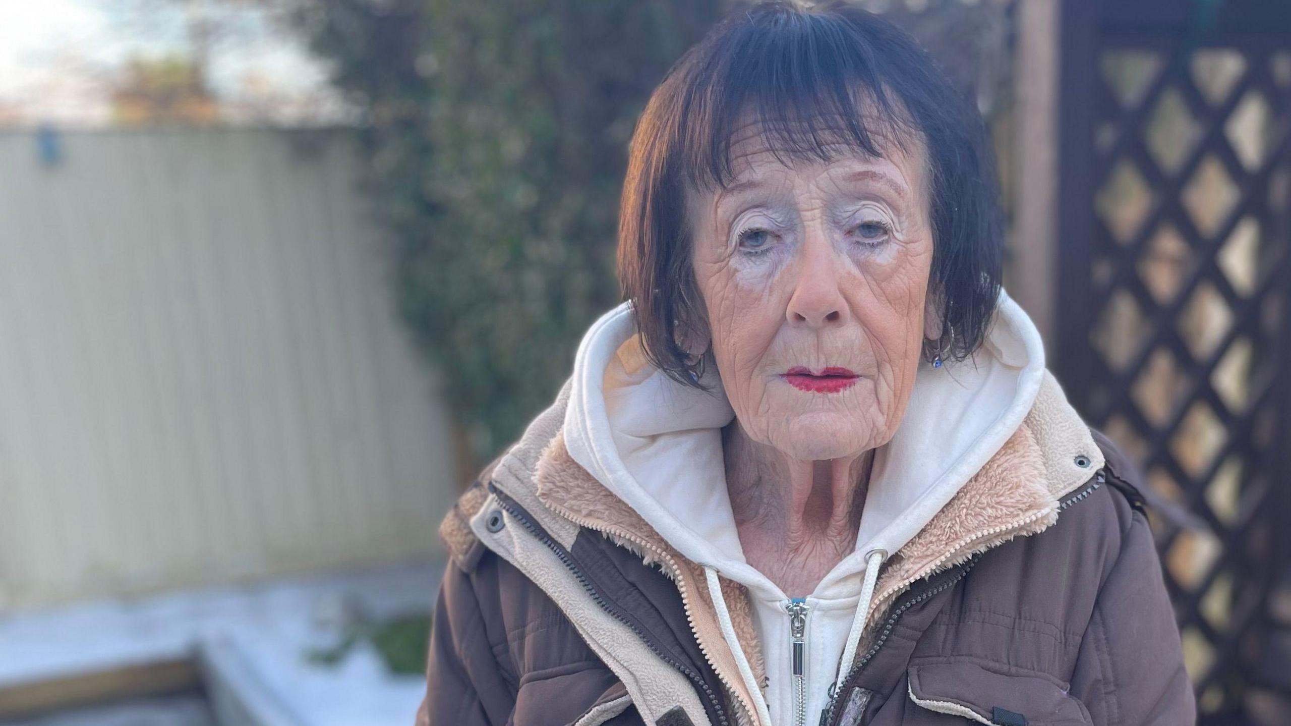 An elderly woman, wearing makeup and a brown coat, with short brown hair, stands outside in her snowy garden, looking at the camera 