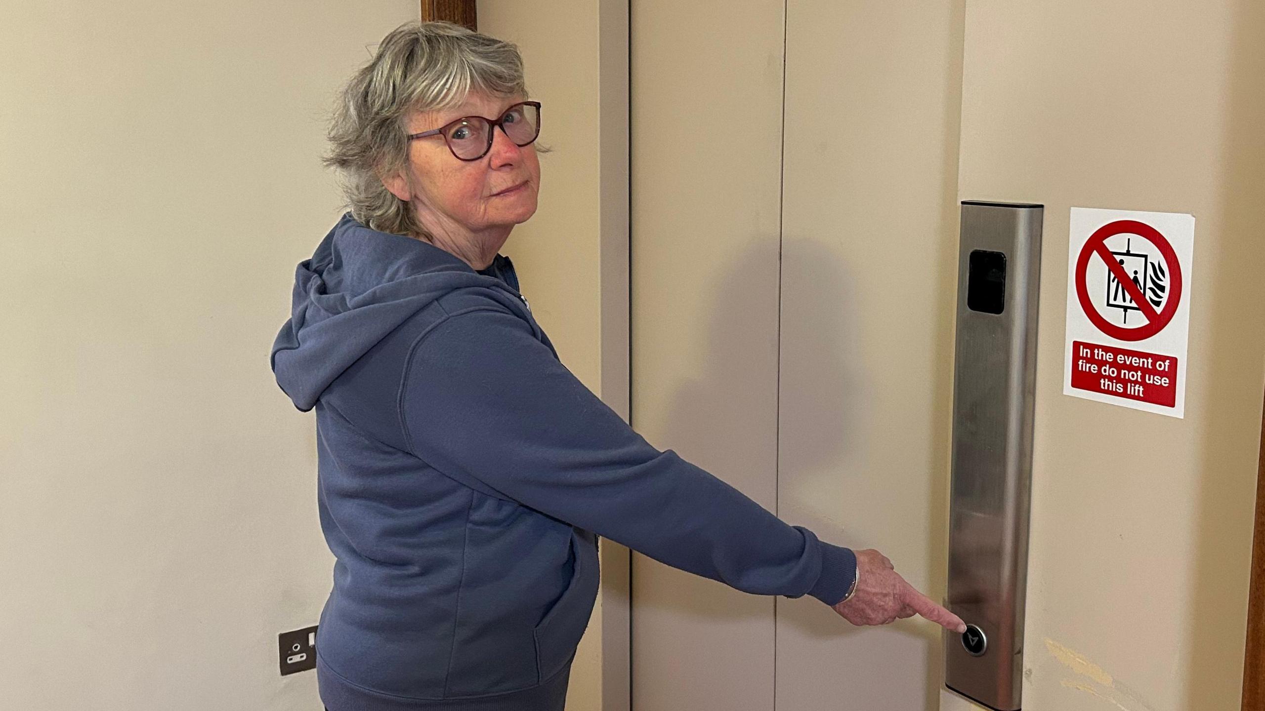 An elderly women standing in front of a closed lift door, pressing the call button. There is a sign on the wall next to the elevator with a red circle and diagonal line which says not to use the lift in the event of a fire. She is wearing a blue jumper and wearing glasses.