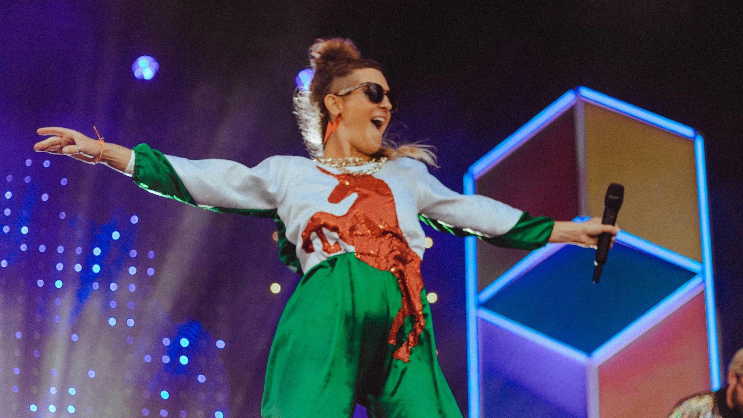 Tara Bandito stands performing, she is wearing a Welsh flag jumpsuit, has her hands outstretched and her hair tied up. She is wearing sunglasses. The background is vibrant blues, purples and reds, with different lights shining on the stage. 