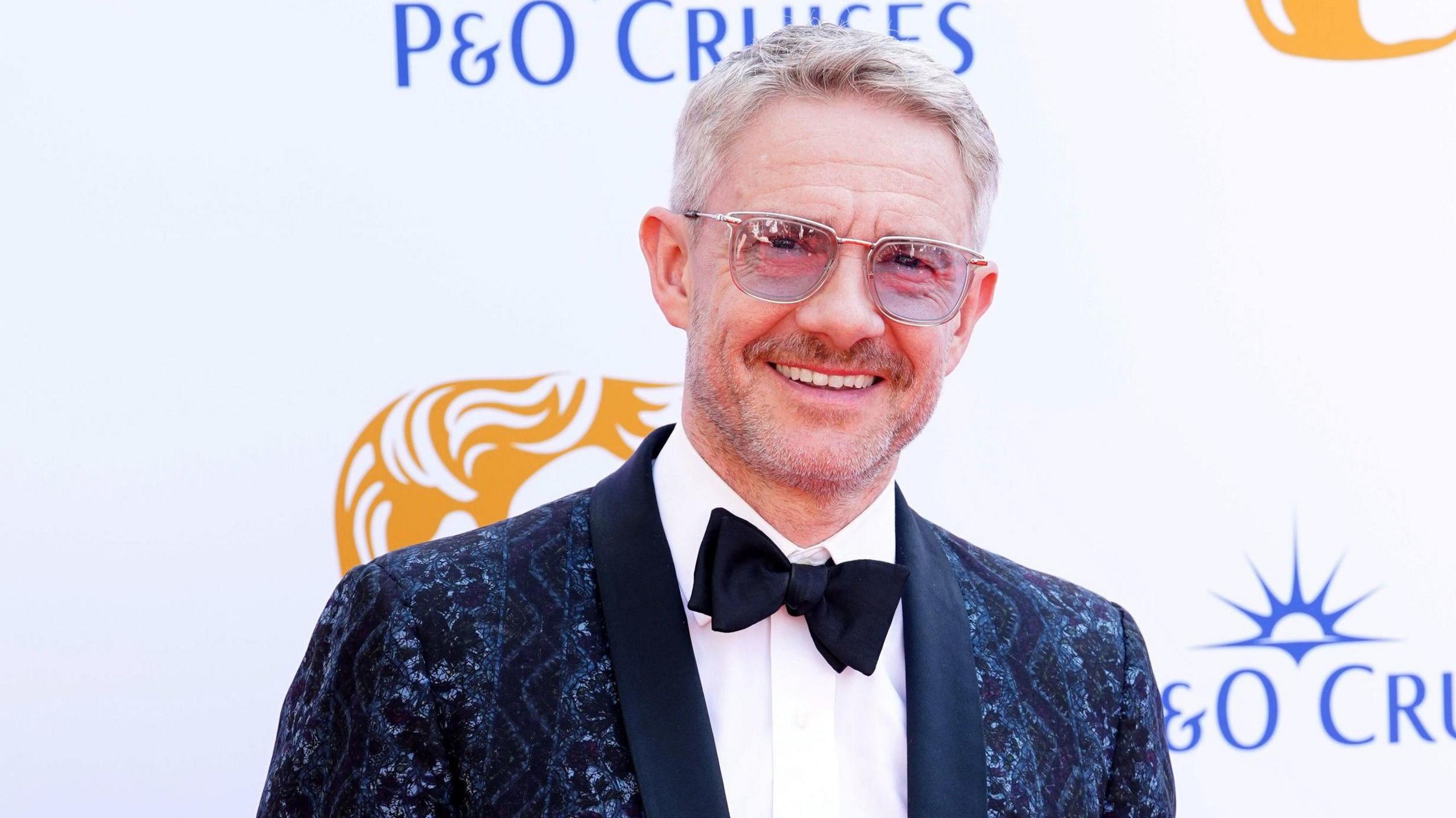 The actor Martin Freeman smiles at the camera. His is wearing a black tie-style outfit and sunglasses. His jacket is dark blue with a paisley pattern worked into it. The lenses of his sunglasses are a very light purple