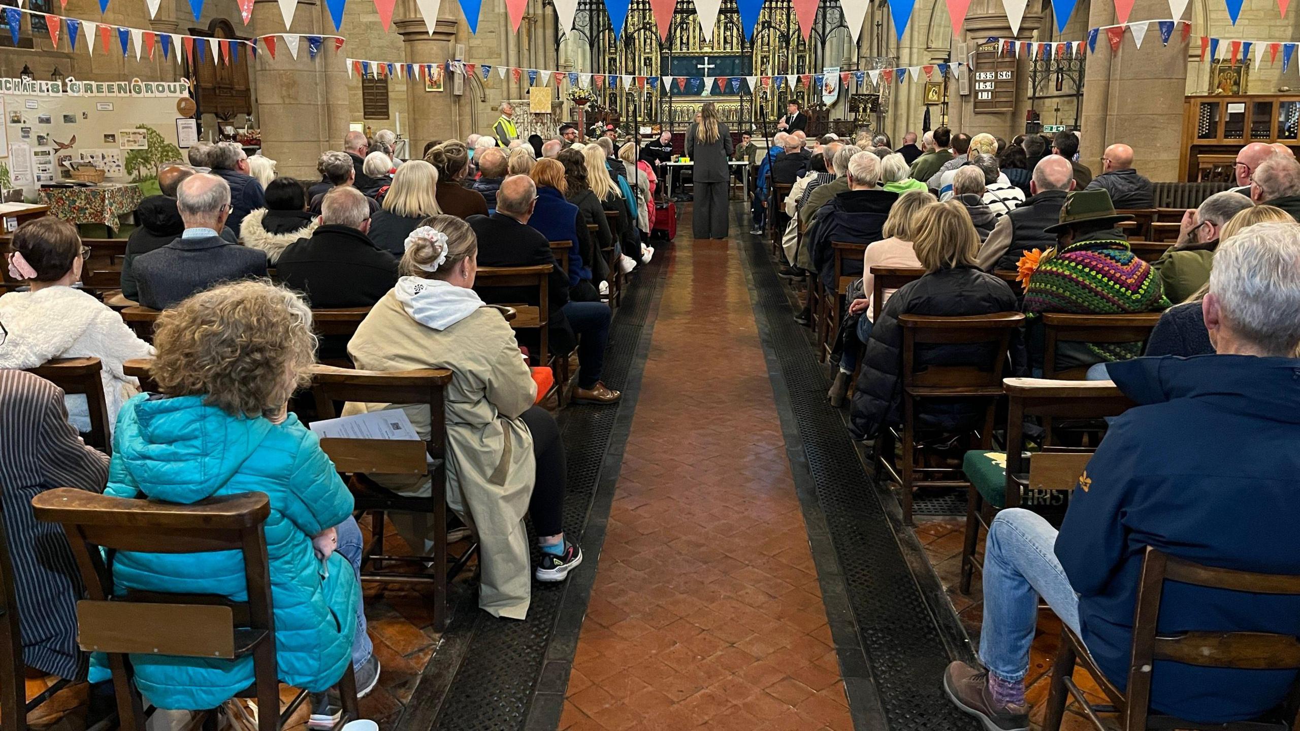 The picture shows the inside of a church, packed with people on every one of the 10 rows on either side. A woman stands at the end of an aisle in front of a long desk which has several people seated on it. Her back faces the crowd. Bunting is attached to the pillars of the church.