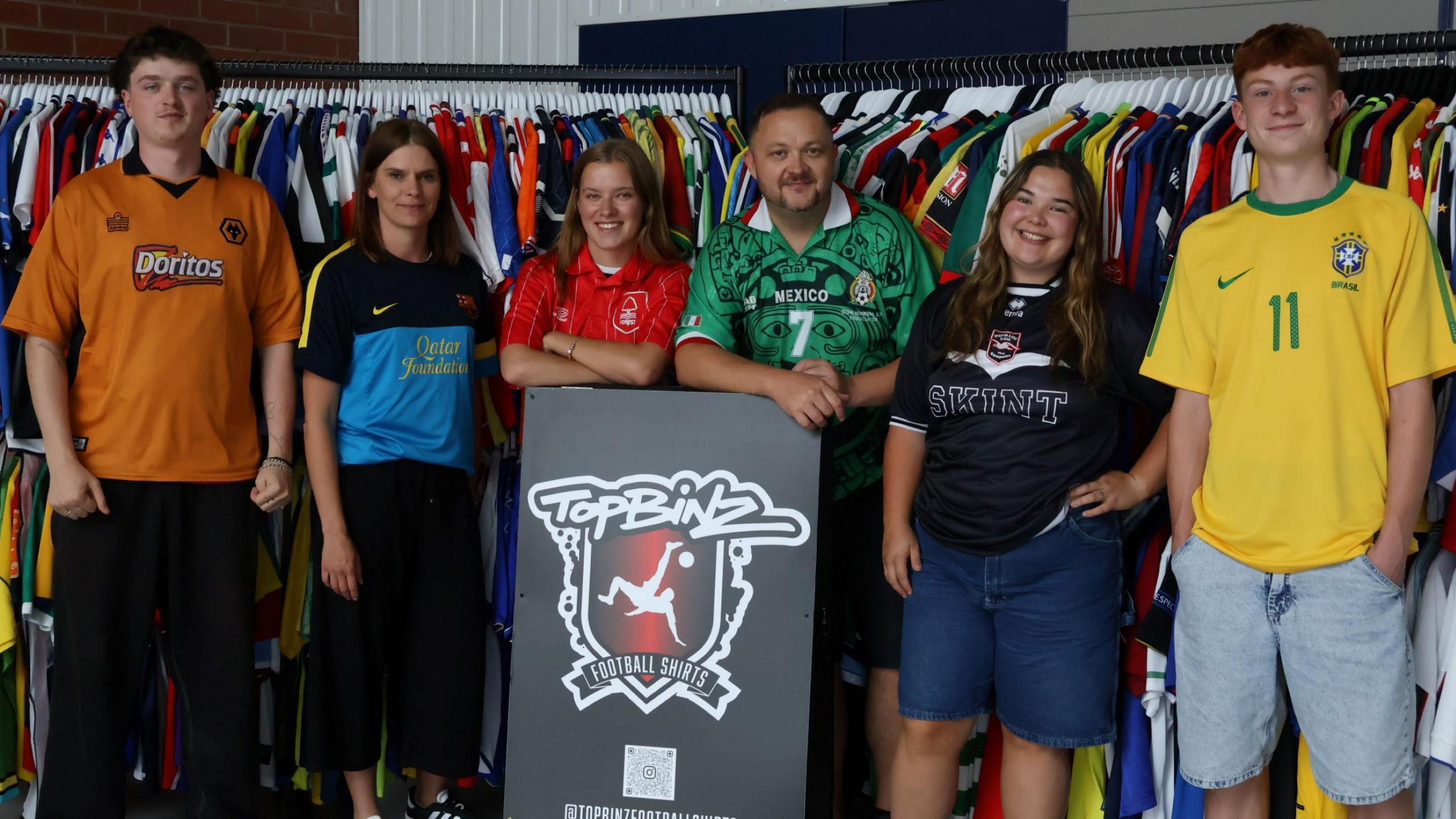Six staff members, three men and three women, stand in front of a row of football shirts on hangers at Bristol vintage football shirt shop TopBinz. From left, they are wearing a Wolves, Barcelona, Nottingham Forest, Mexico, Brighton and Brazil top.