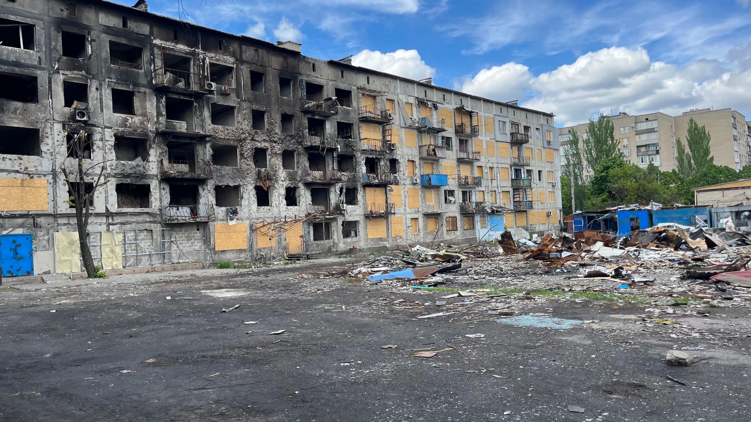 A long five-story building has scorch marks across it with many windows missing. Some of the holes have been boarded up. A large pile of debris lies on tarmac in front of the building.