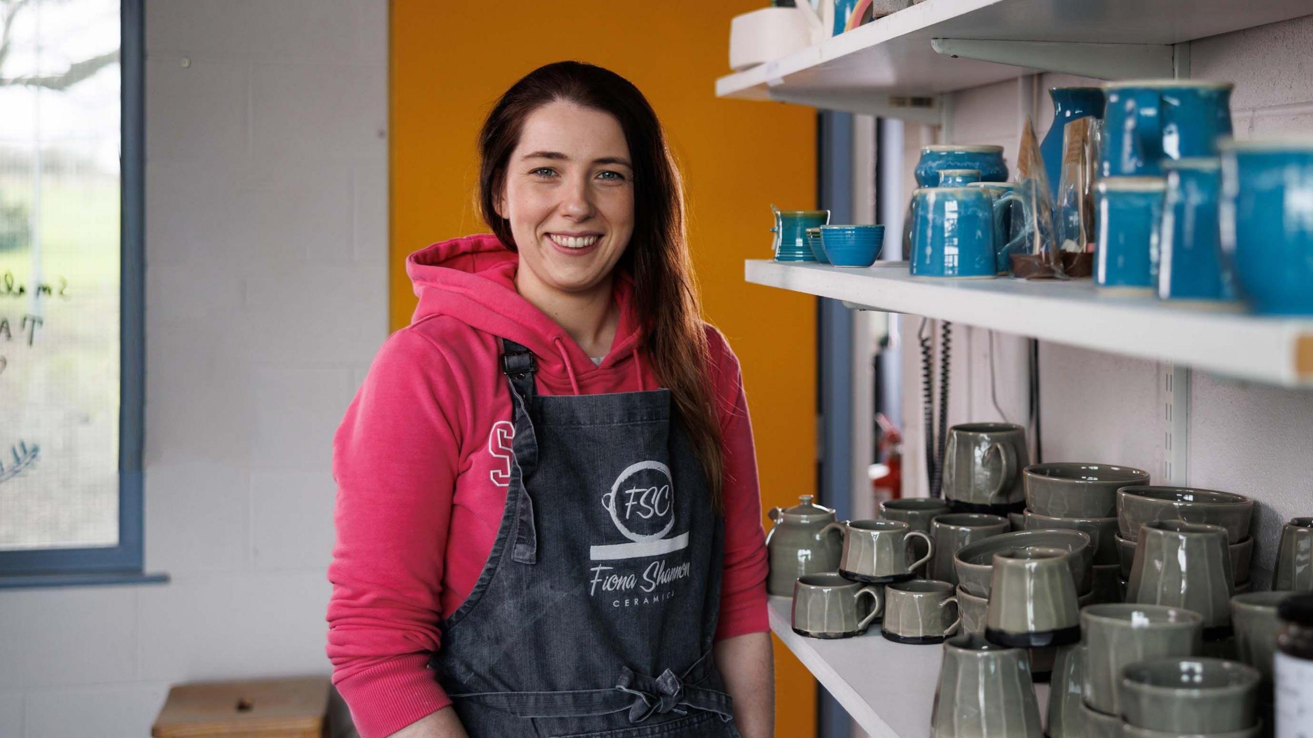 Fiona Shannon - a woman with long, dark hair wearing a pink hoodie and dark apron smiles at the camera. She is standing beside a shelf of blue and green mugs.