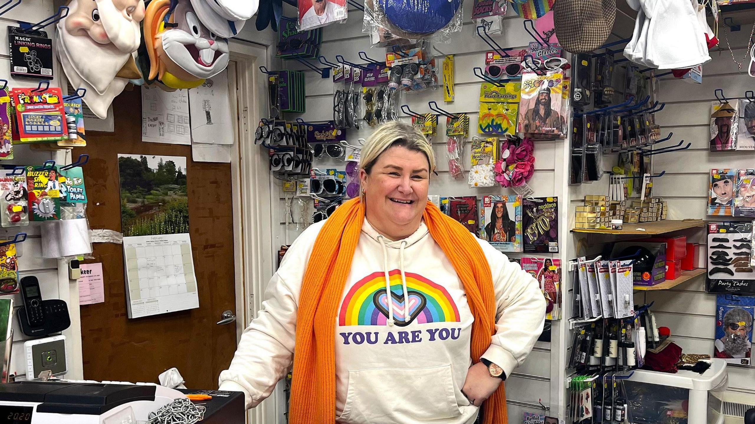 Sara Hennessy in a white hoodie and a orange scarf standing behind the counter of a shop with costume items in the background.