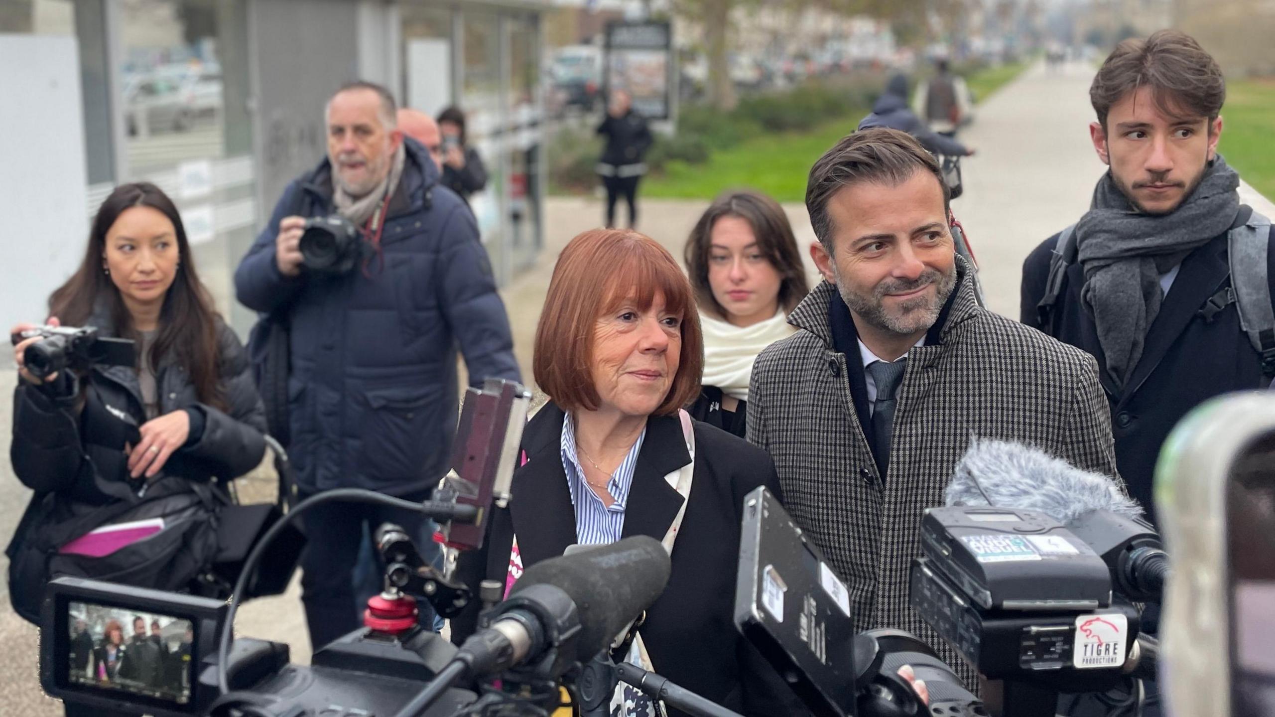 Gisèle Pelicot is surrounding by members of the press carrying cameras. She is walking with her head held high with a faint smile. She has a red bob and is wearing a black coat. She is flanked by her legal team.