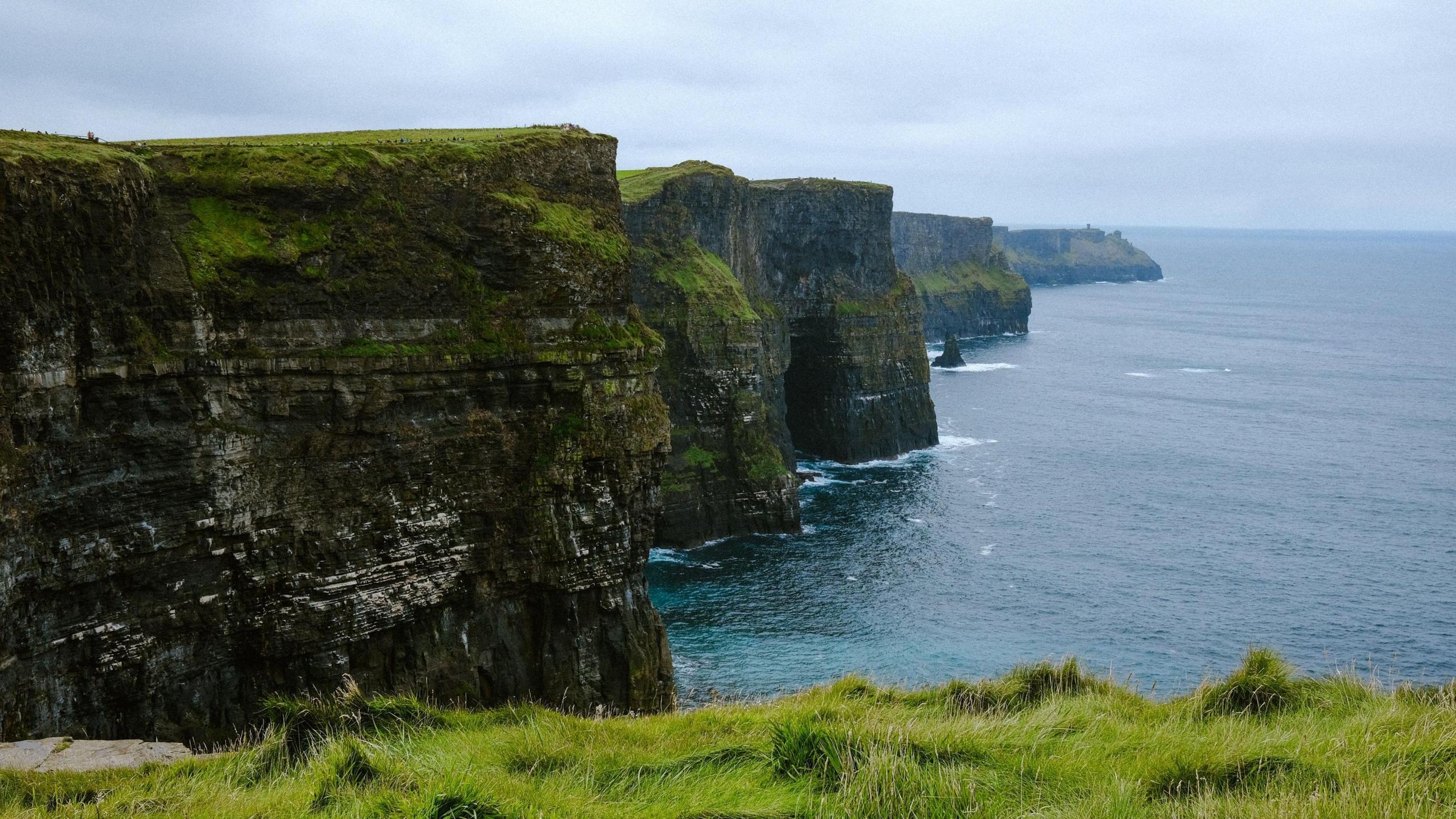 A view of the Cliffs of Moher on a cloudy day with a calm sea below. The tall, rugged cliffs jut out at angles to the sea.