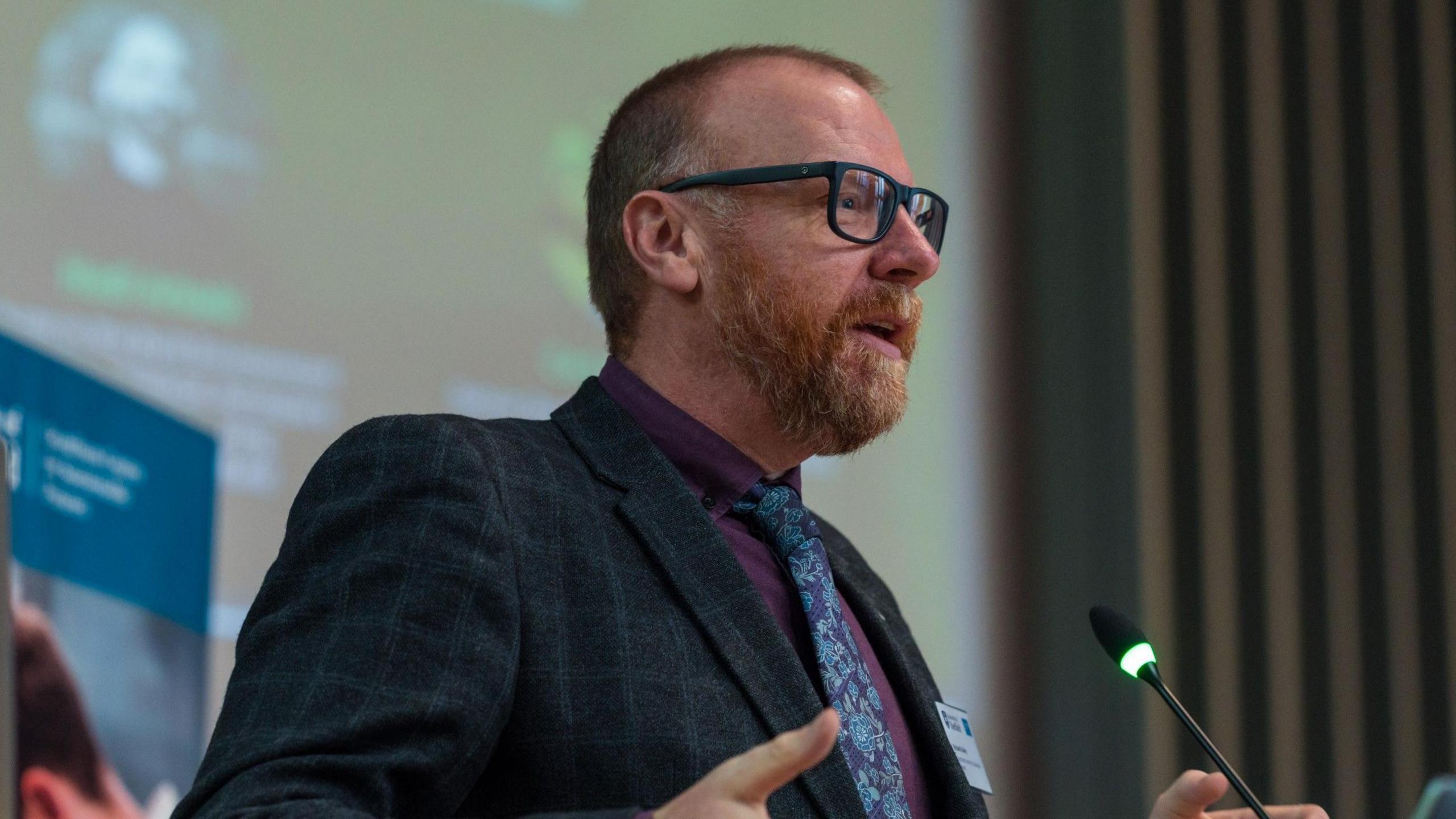 A man with black rimmed glasses and a beard is wearing a dark suit with a purple shirt and blue patterned tie. He is stood in front of a microphone with his hands slightly raised giving a speech