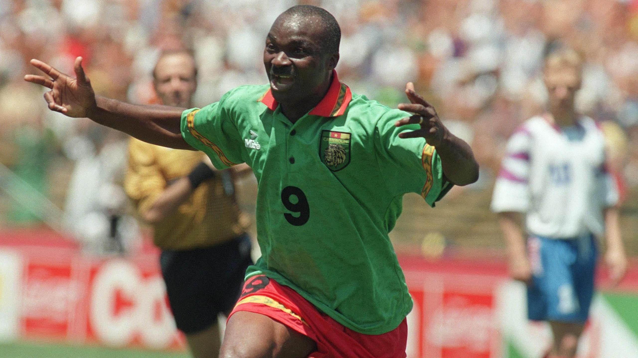 Roger Milla in green Cameroon jersey wheels away with both arms raised in celebration after scoring a goal at the 1994 World Cup with a forlorn-looking Russia defender and the referee in the background.