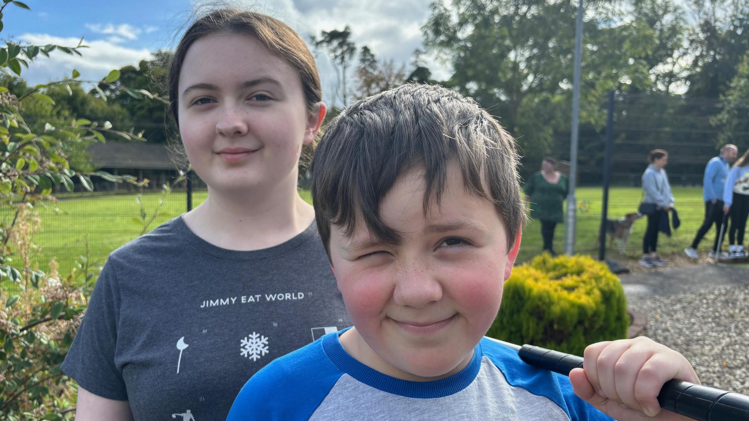 Ellie and Zack standing at the miniature golf course. Ellie is wearing a grey t-shirt and has her dark hair pulled back in a ponytail. Zack has short dark hair and is wearing a blue and grey top. He is squinting because of the sun and is carrying a putter in one hand