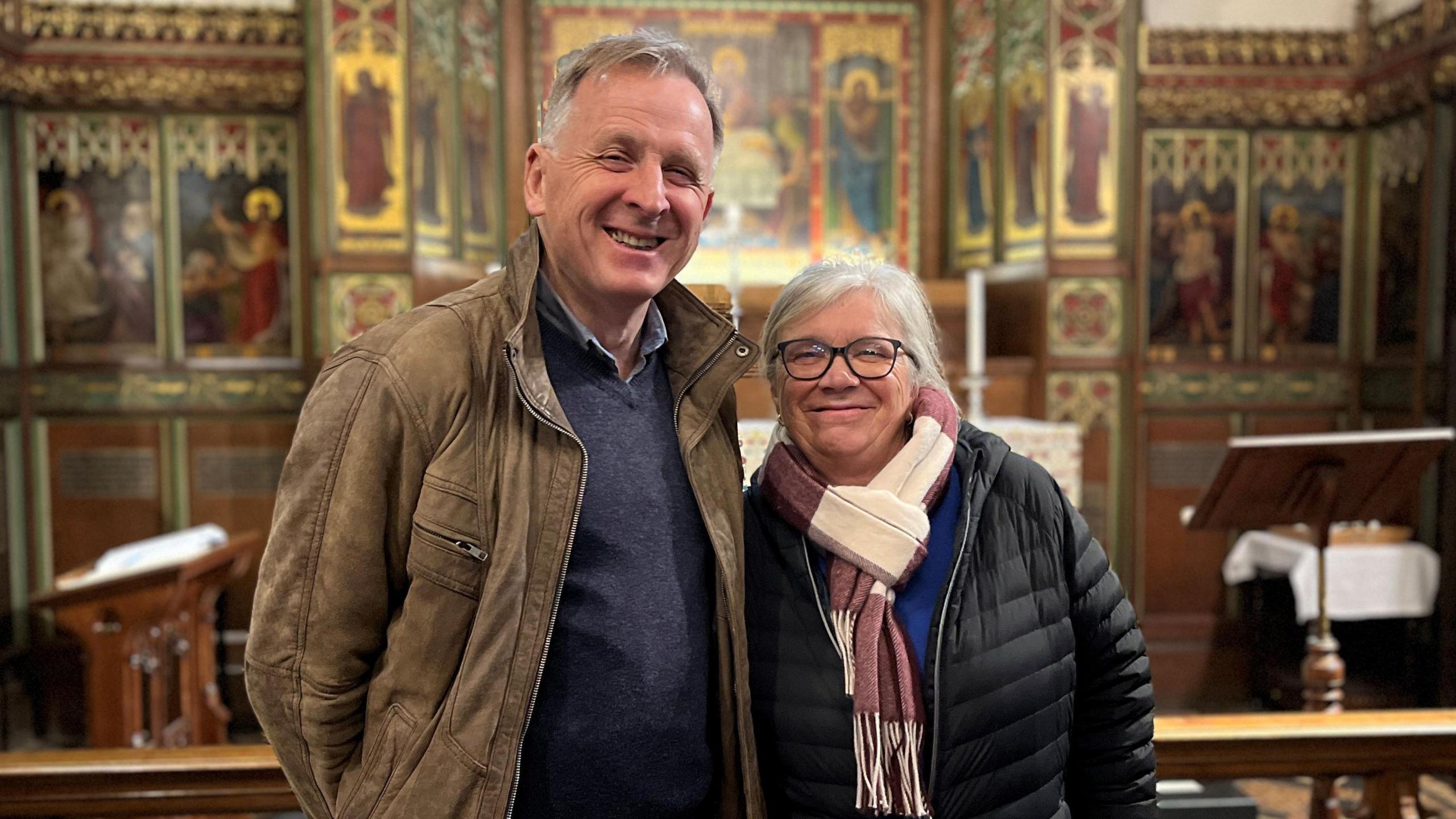 David Matthews and Jo Jones standing together inside St Mary Le Tower church, smiling at the camera. David has short grey hair and is wearing a navy jumper and beige coat. Jo has shoulder-length grey hair and is wearing glasses. She has a royal blue top on, with a black puffer jacket and pink and white scarf over the top. The church's altar is behind them.