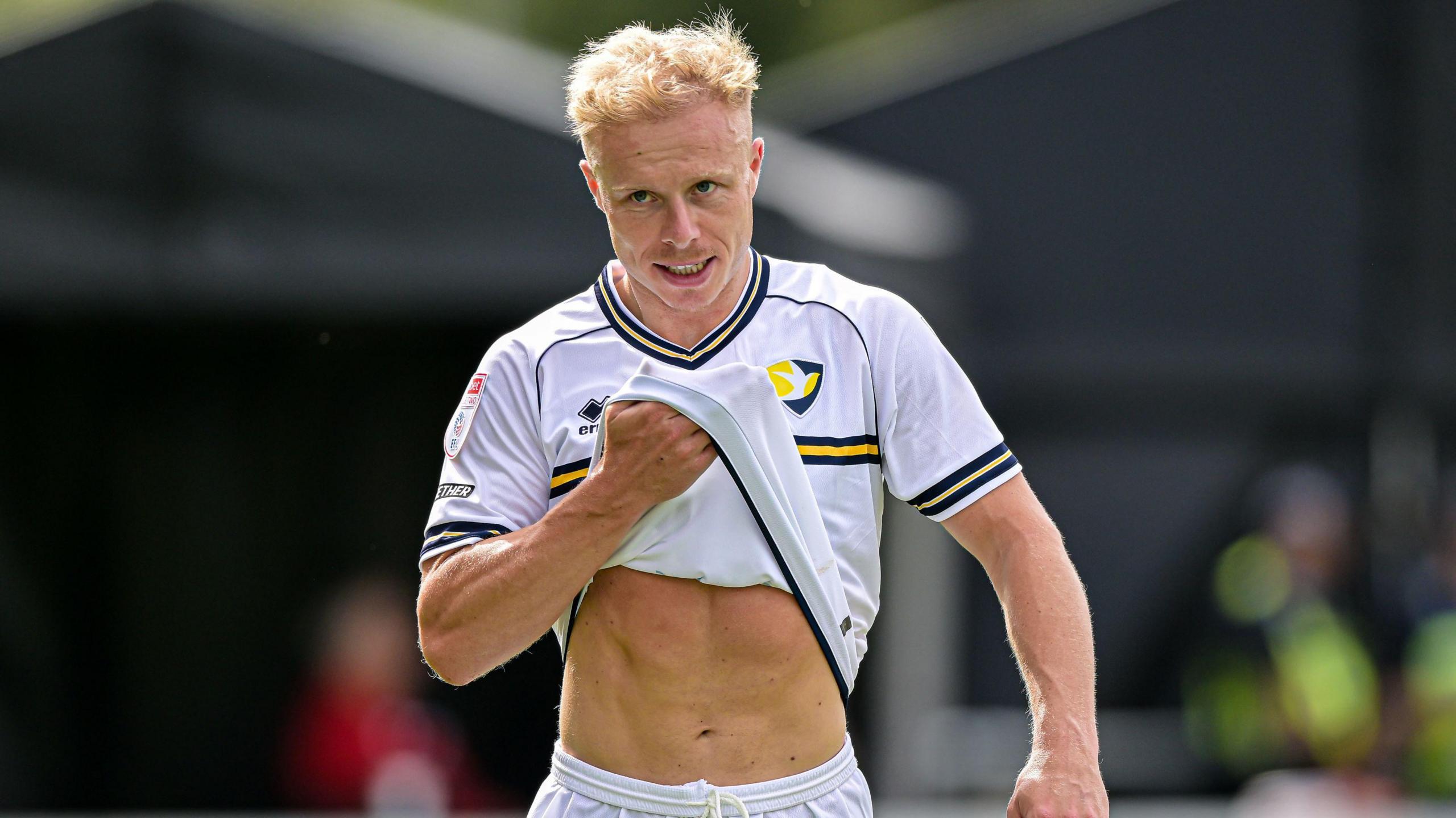 Cheltenham Town player Ryan Broom wipes his face with his white shirt as he is substituted during the match with Cambridge United. He has short blonde hair.
