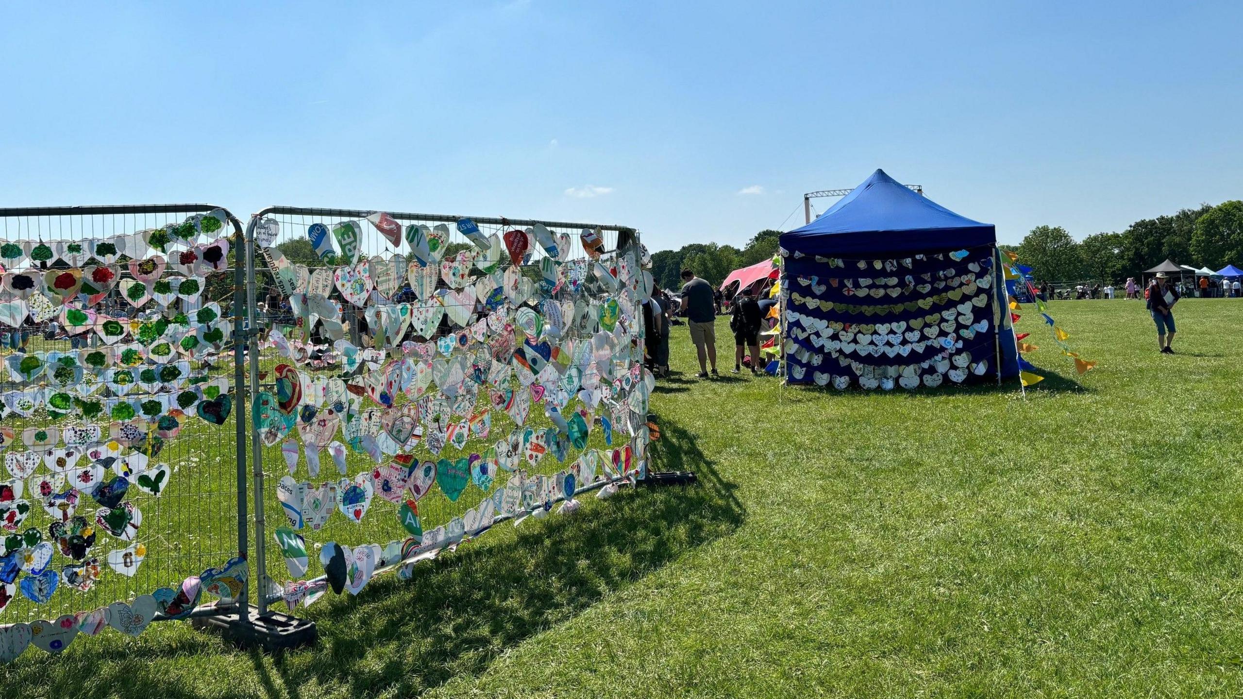 Grilled frame with thousands of paper hearts placed on green grass - and a blue tent with more rows of paper hearts displayed on it.