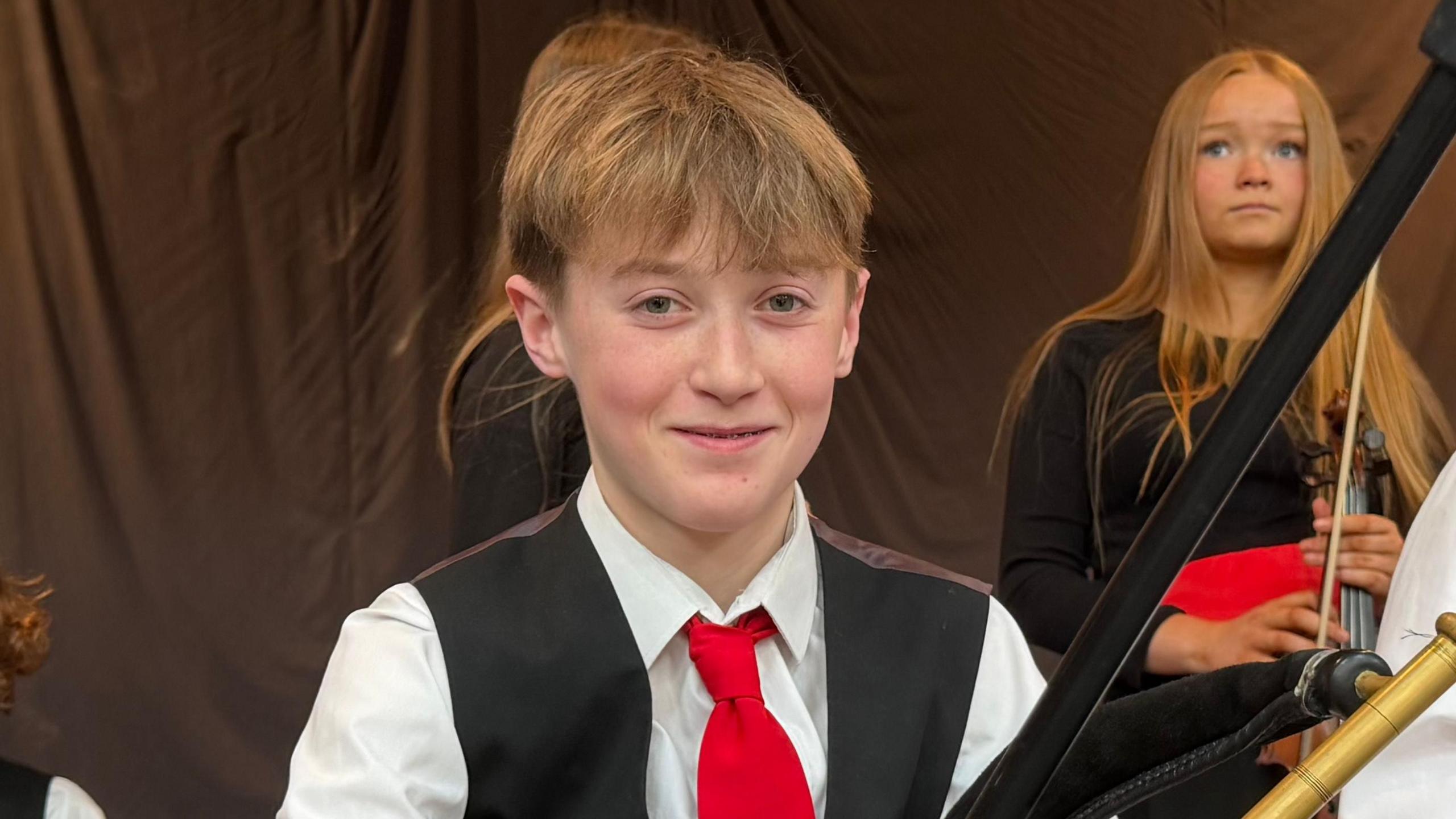 A boy with short blonde hair, wearing a white shirt, red tie and black waistcoat. There is a boy and a girl sat behind him. He is holding uilleann pipes.