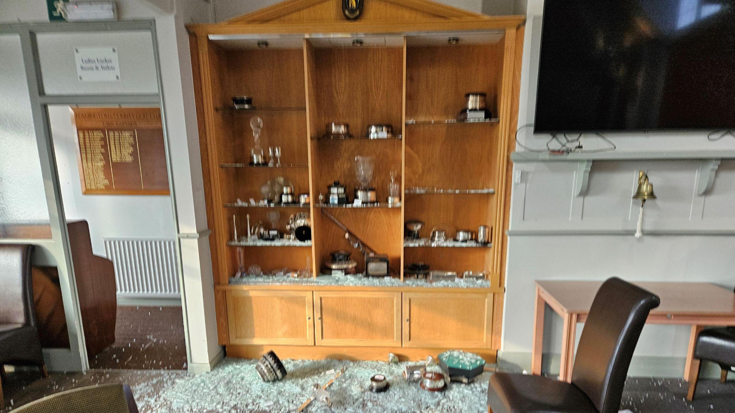 A smashed cabinet with the remains of a number of trophies on the floor, surrounded by broken glass